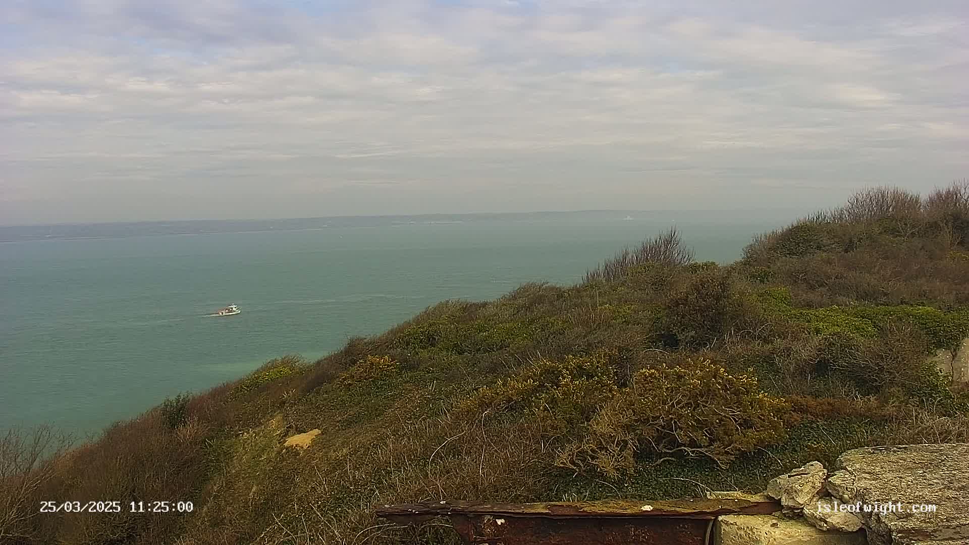A tranquil coastal scene under a cloudy sky shows a calm sea extending to the horizon, with a small boat visible in the distance, and a hillside covered in low-lying vegetation in the foreground.