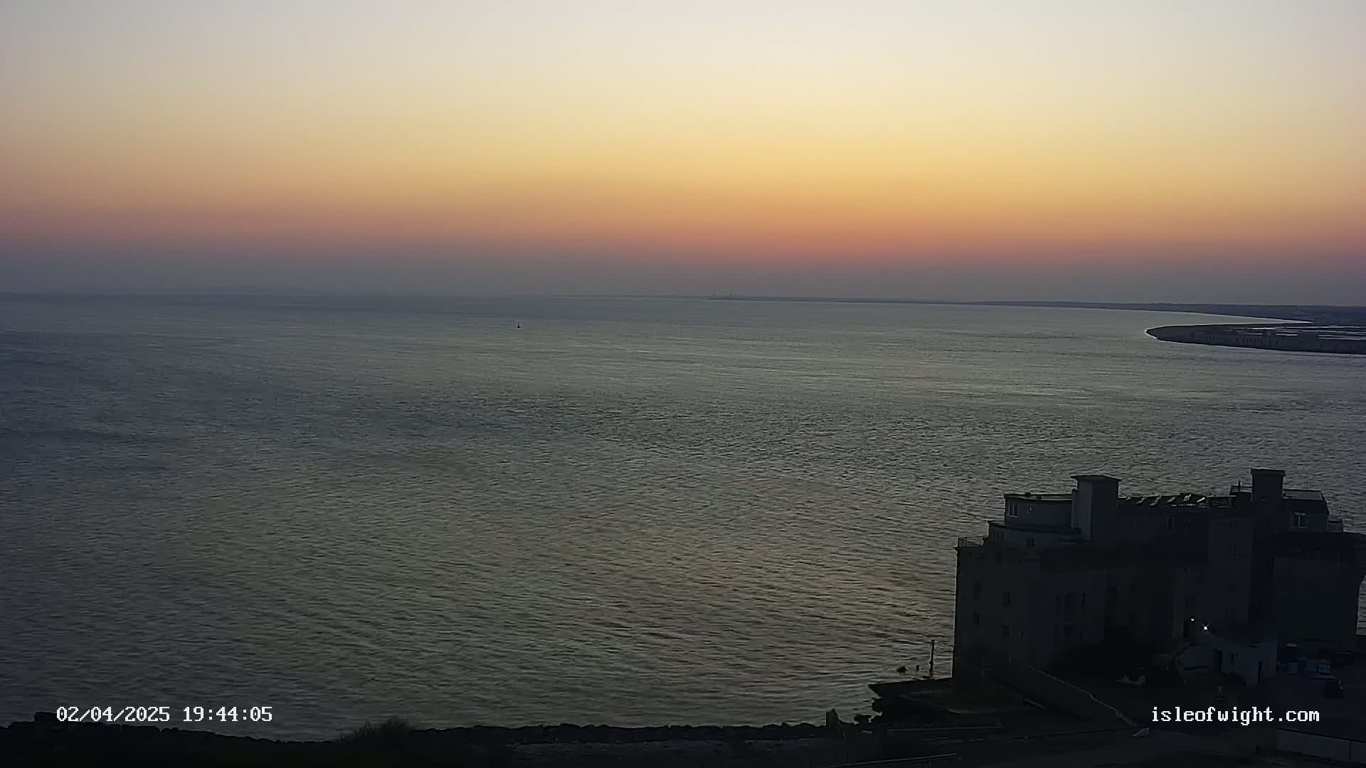 A large body of water under a sunset sky is viewed from above, with a dark building situated on the lower right.