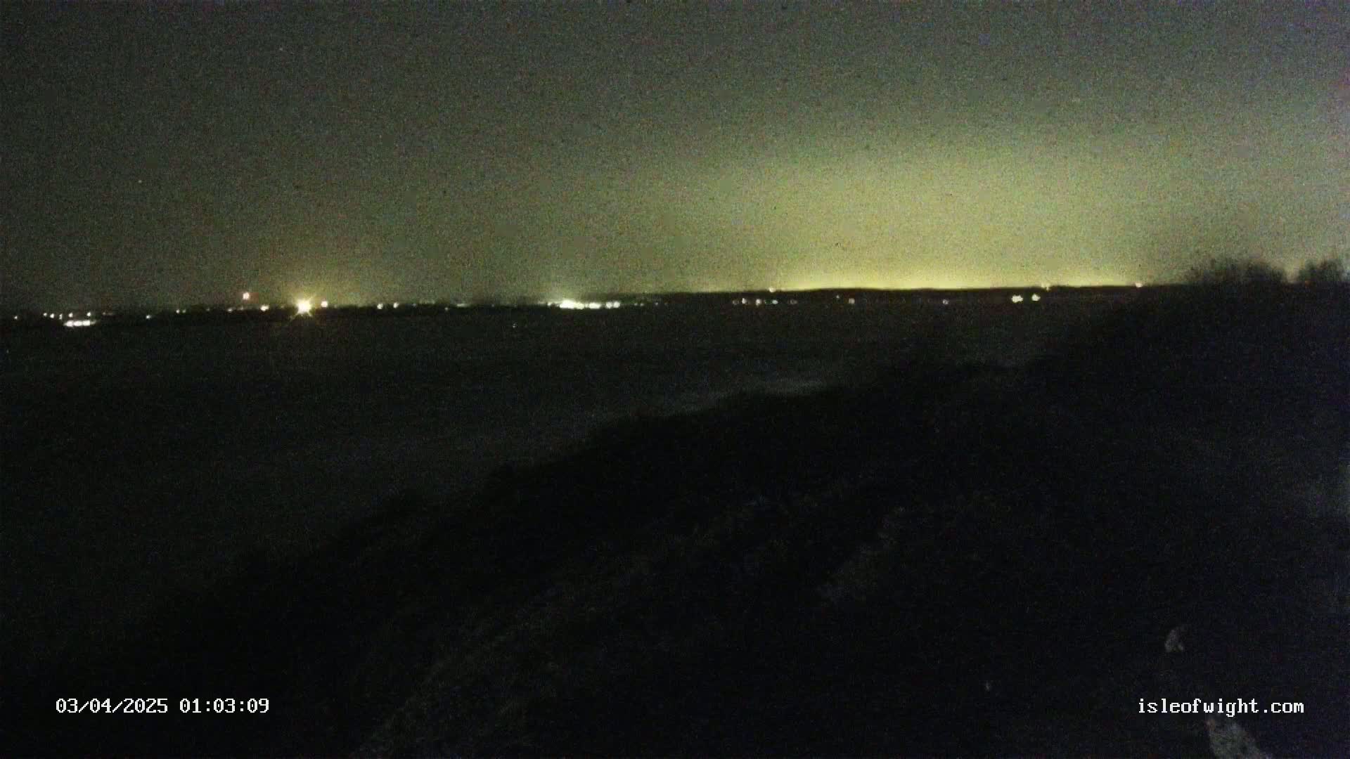 A nighttime view of a dark body of water with a faintly illuminated shoreline in the distance under a dark, possibly overcast sky.