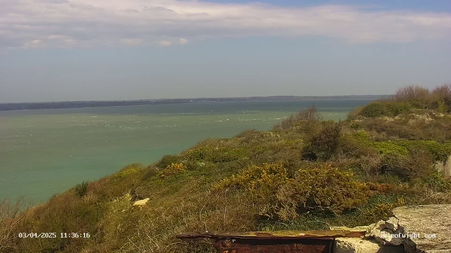 A view from a hillside shows a calm, green-tinged body of water under a partly cloudy sky.