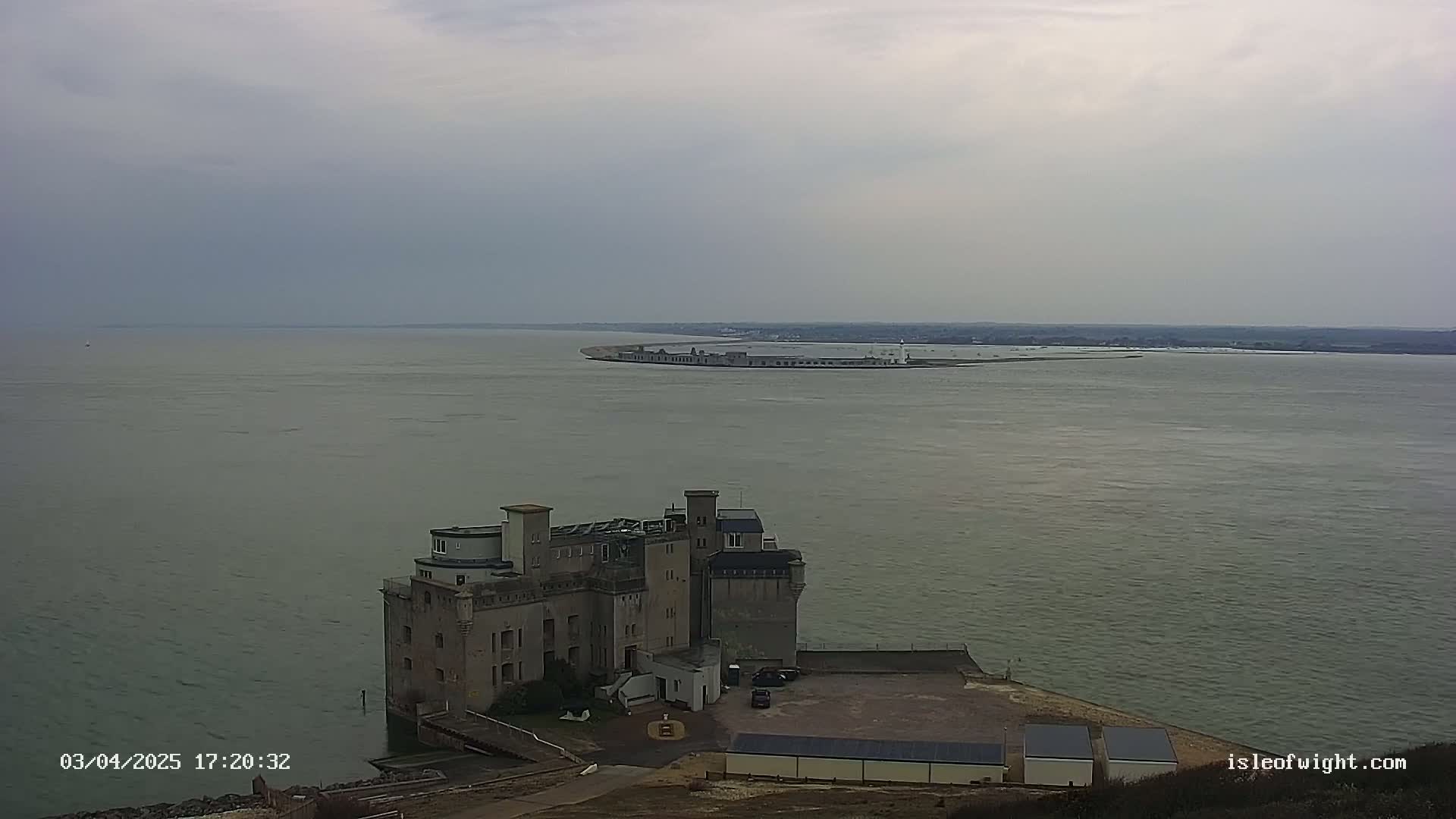 A stone castle-like structure sits on a cliff overlooking a calm body of water, with a long, low structure visible in the distance under an overcast sky.