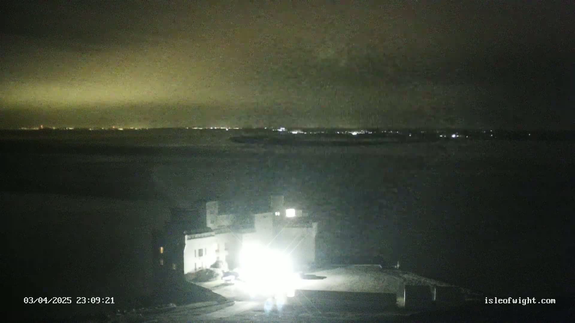 A brightly lit building is in the foreground, with a dark body of water and a distant, faintly lit cityscape visible under a dark, cloudy night sky.
