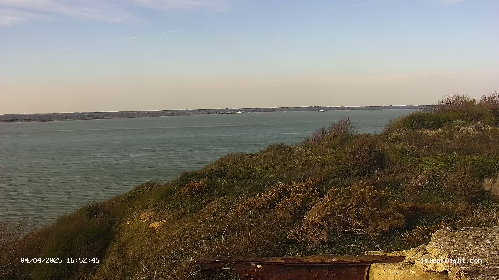 A calm, wide body of water is seen from a hillside covered in low-lying shrubs and vegetation under a mostly clear sky.