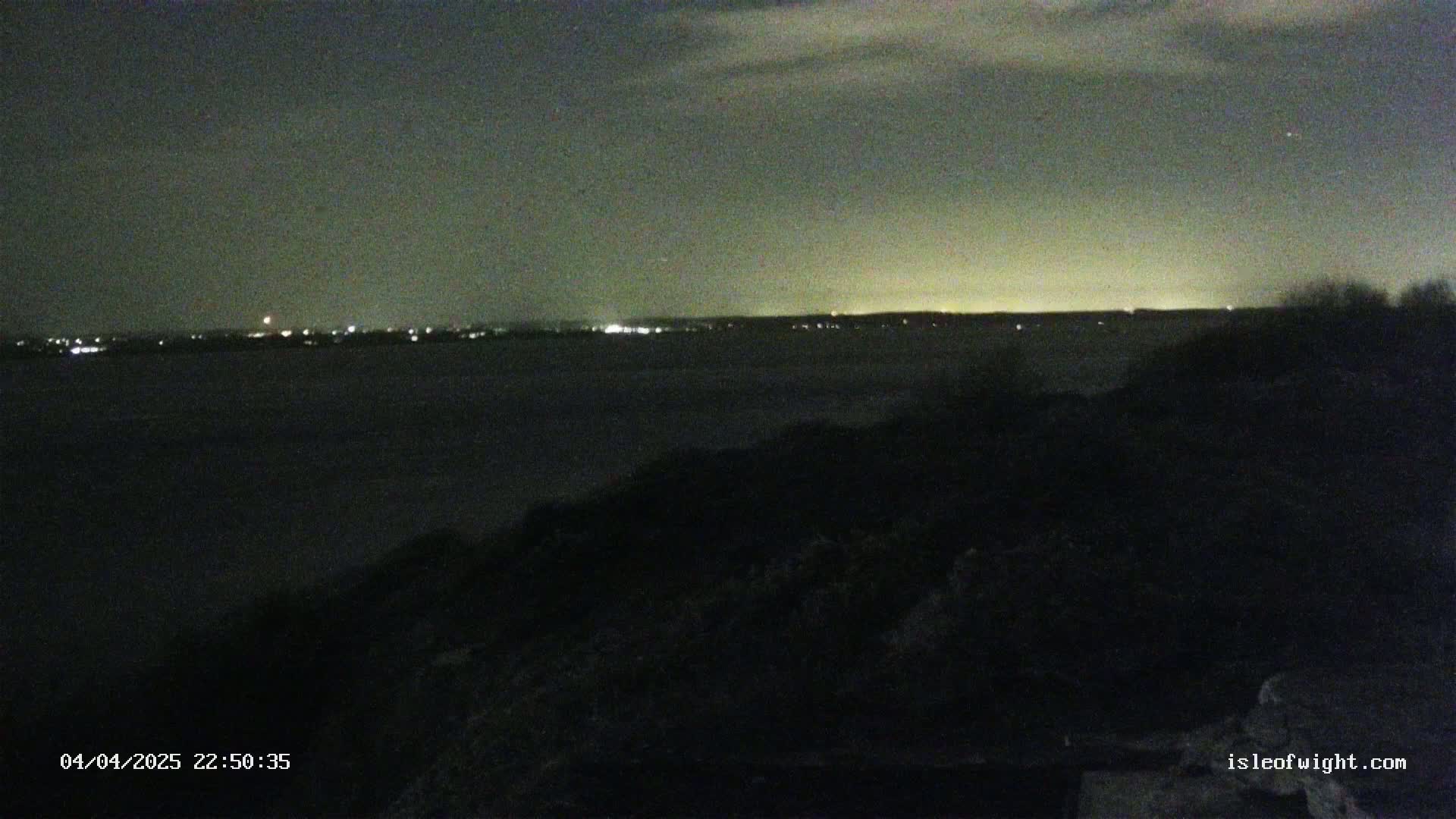 A nighttime view shows a dark body of water with a distant, faintly lit shoreline under a cloudy sky.
