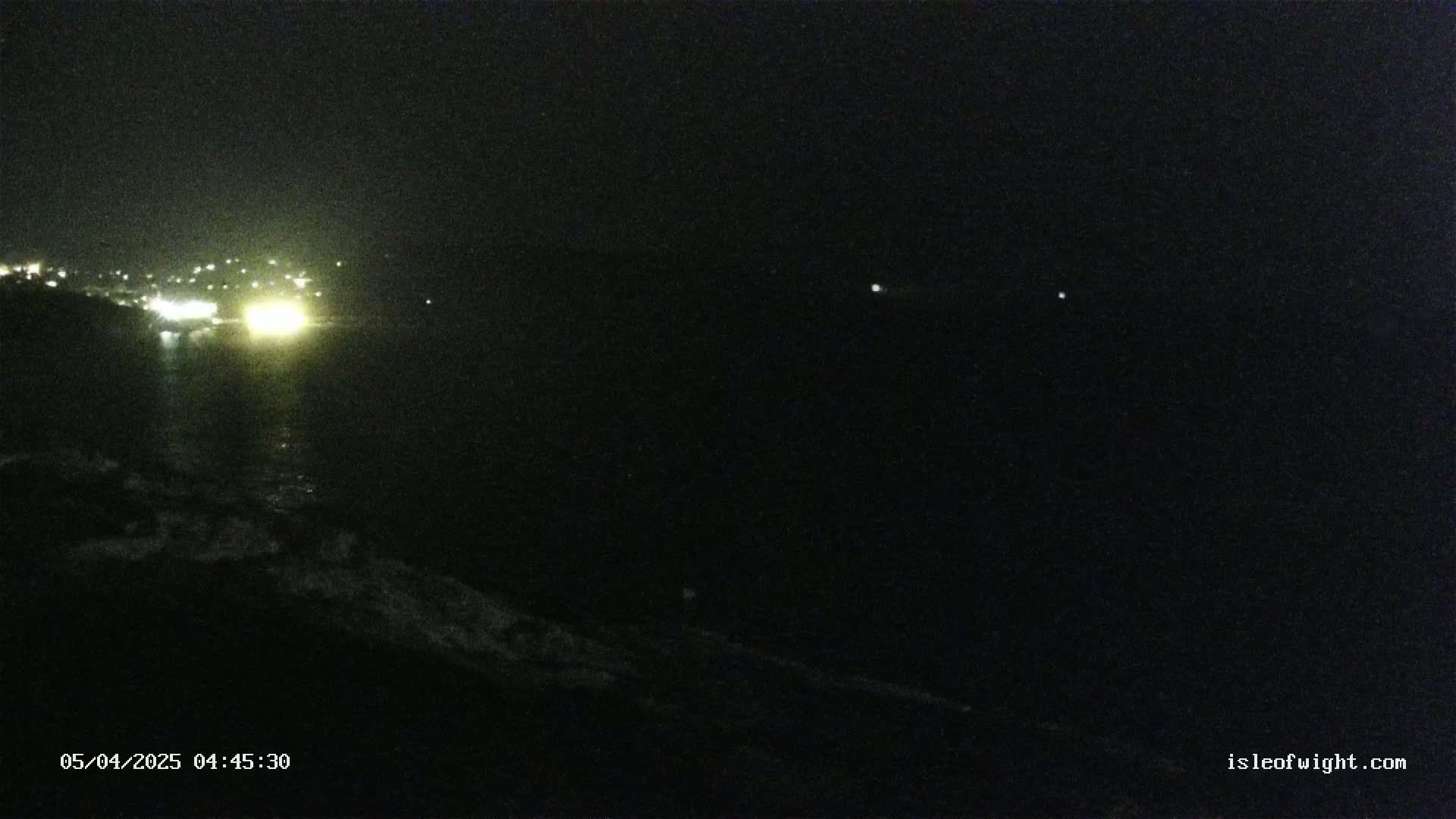 A nighttime coastal scene shows a dark body of water reflecting the lights of a distant town under a clear, dark sky.