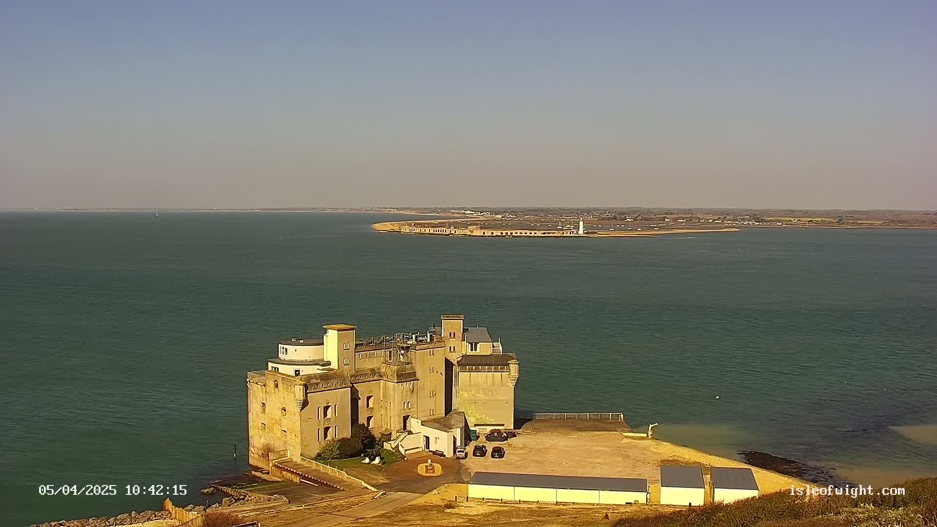 A large, castle-like structure sits on a coastal point overlooking a calm body of water under a clear, sunny sky.