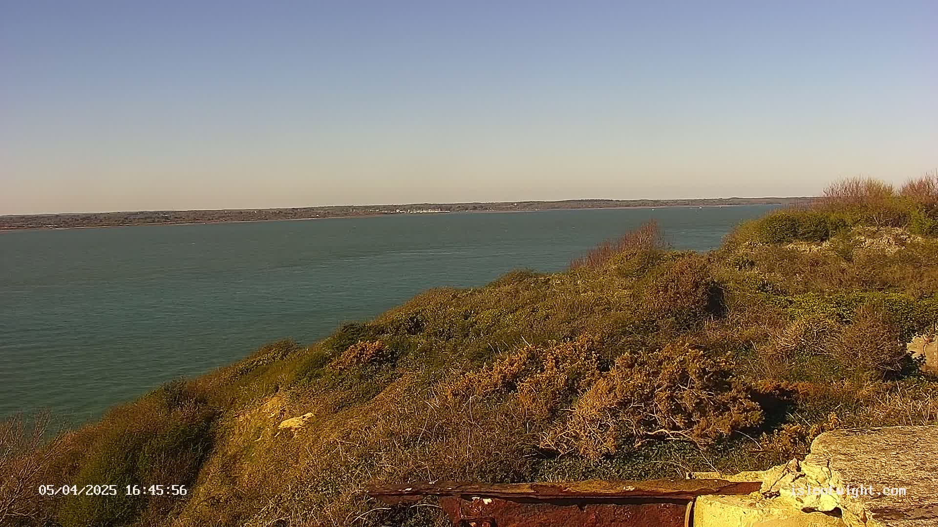 A calm, wide body of water is seen from a high vantage point overlooking a hillside covered in low-lying, scrubby vegetation under a clear, sunny sky.