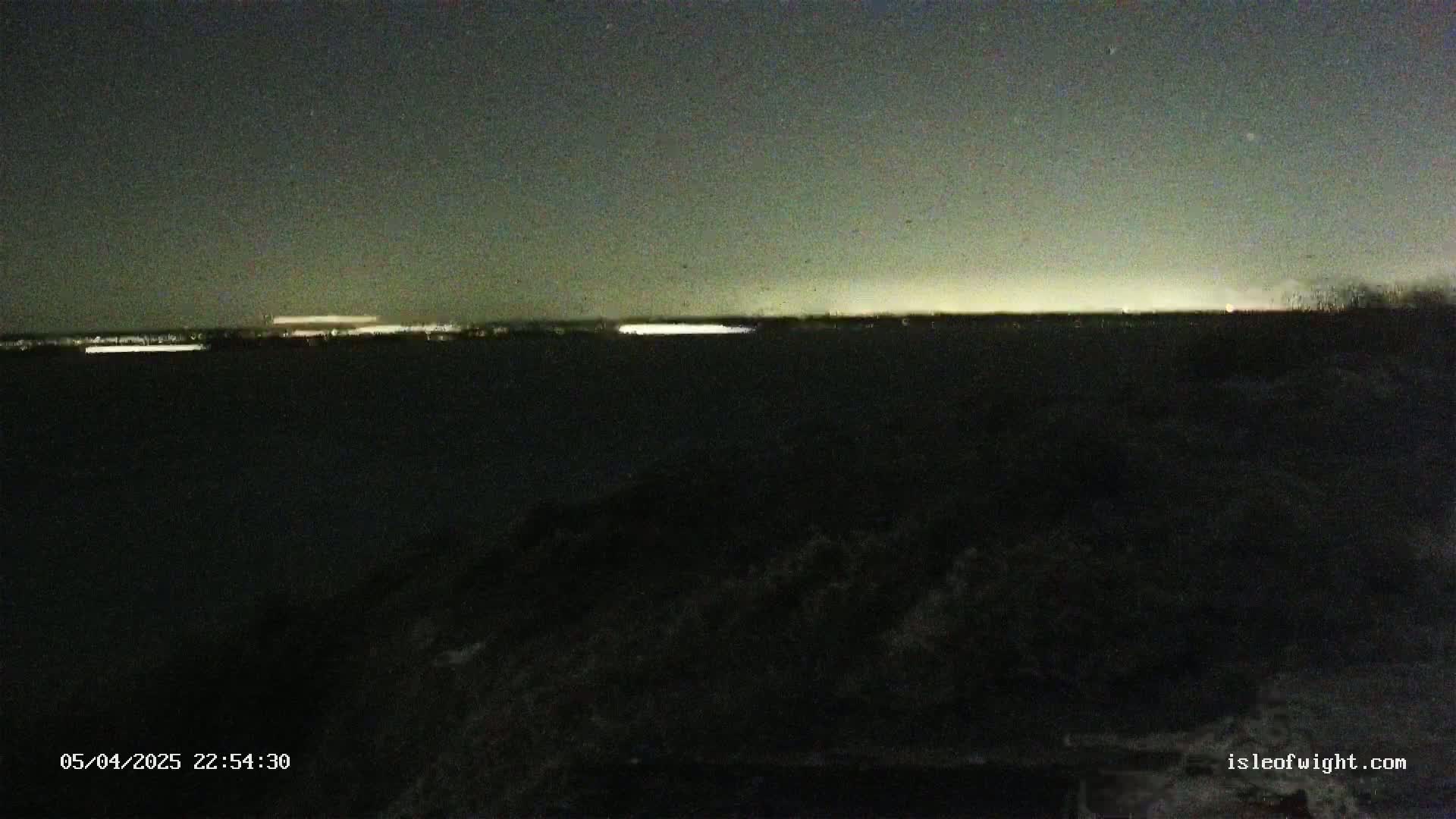 At night, a dark landscape shows a distant horizon with faint city lights under a mostly clear sky.