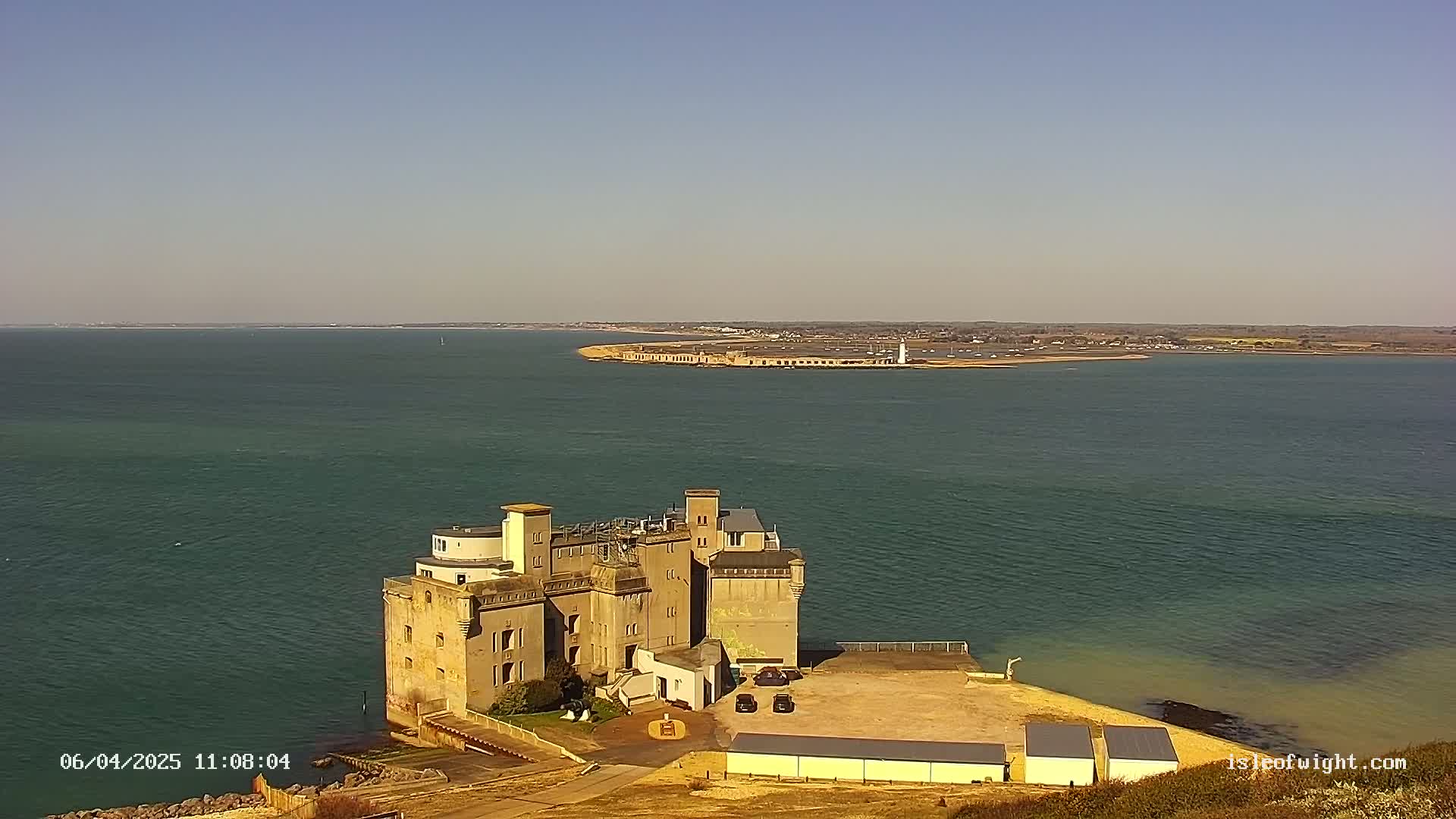 A castle-like structure sits on a coastal cliff overlooking a calm body of water under a clear sky, with a distant landmass visible in the background.
