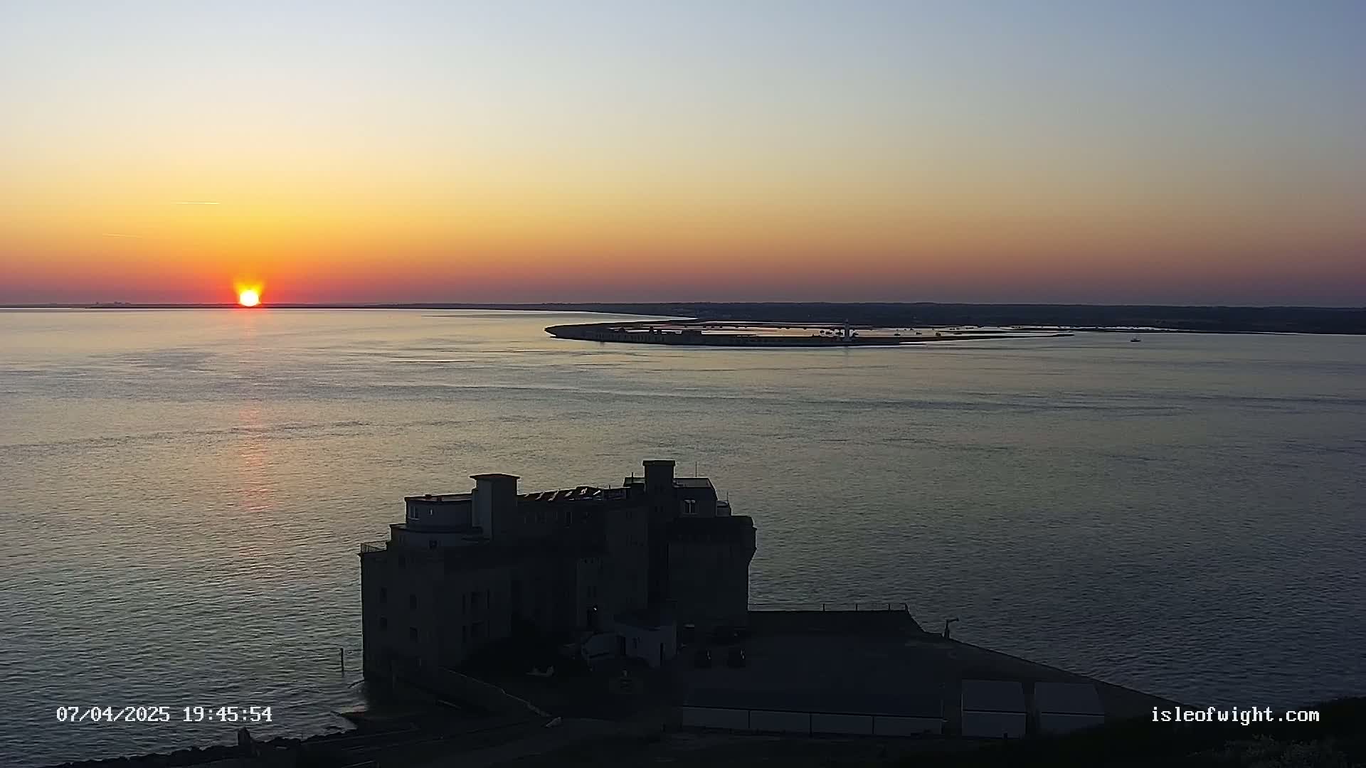 A large, dark building sits on the coast at sunset over calm water, with a distant island visible across the bay.