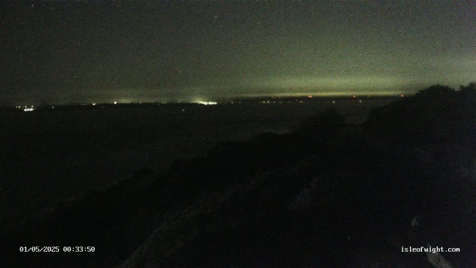 A dark, nighttime view shows a distant, faintly lit shoreline across a body of water, partially obscured by a dark foreground of land.