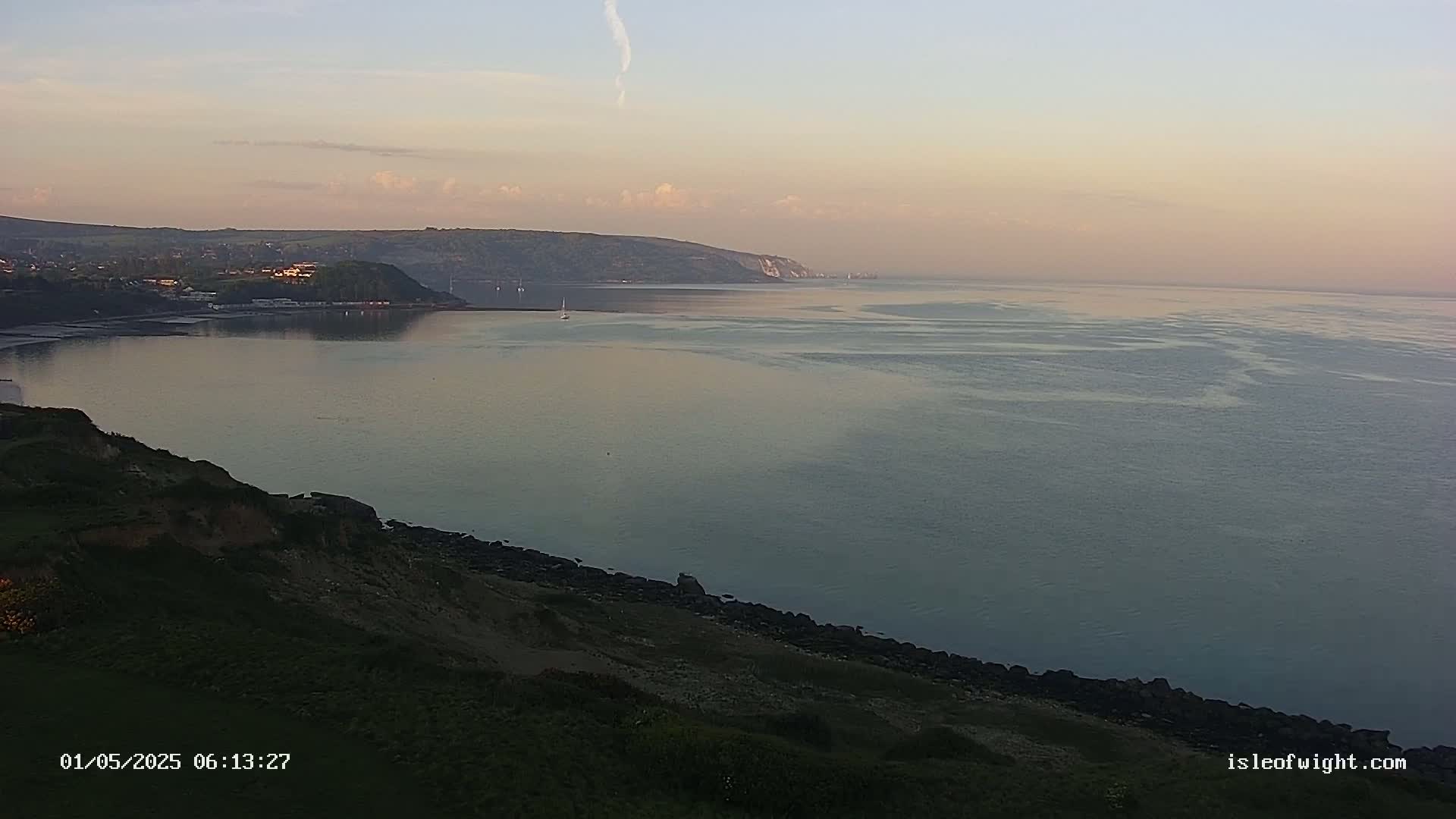 A tranquil coastal scene at dawn shows a calm body of water stretching to a distant, hilly shoreline under a pale sky.
