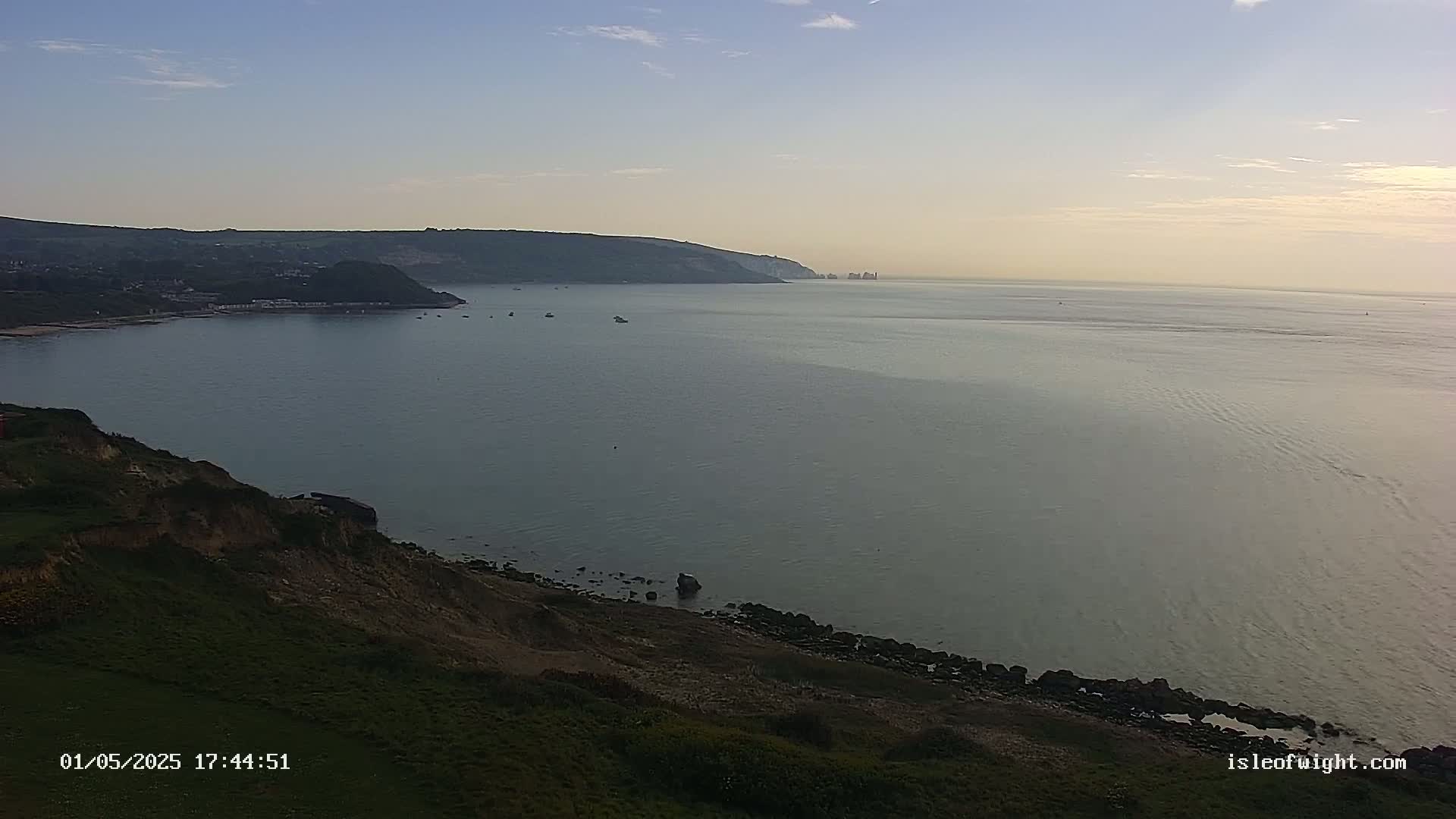 A calm sea stretches out to the horizon under a clear, pale sky, with a grassy cliff and distant coastline visible in the foreground.