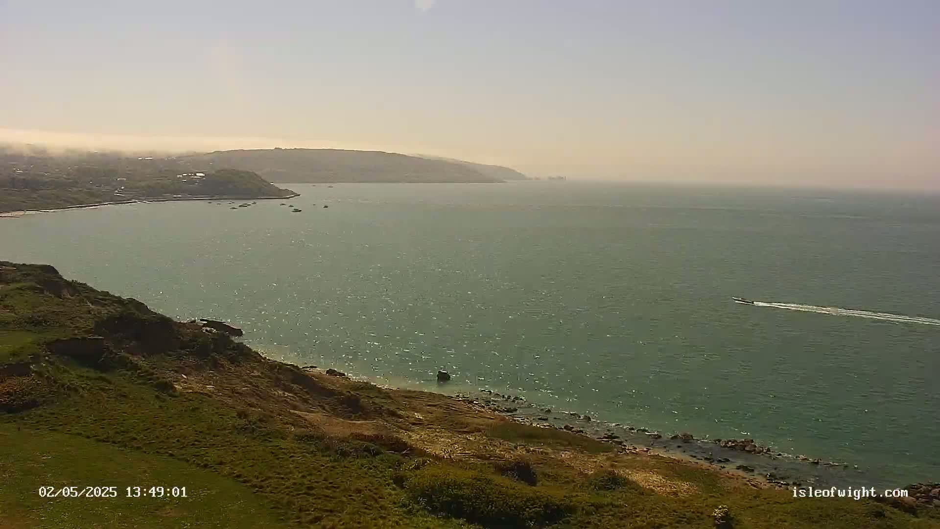 A hazy coastal view shows a green, gently rolling grassy hillside descending to a rocky shore and calm, greenish-blue sea with a few boats.