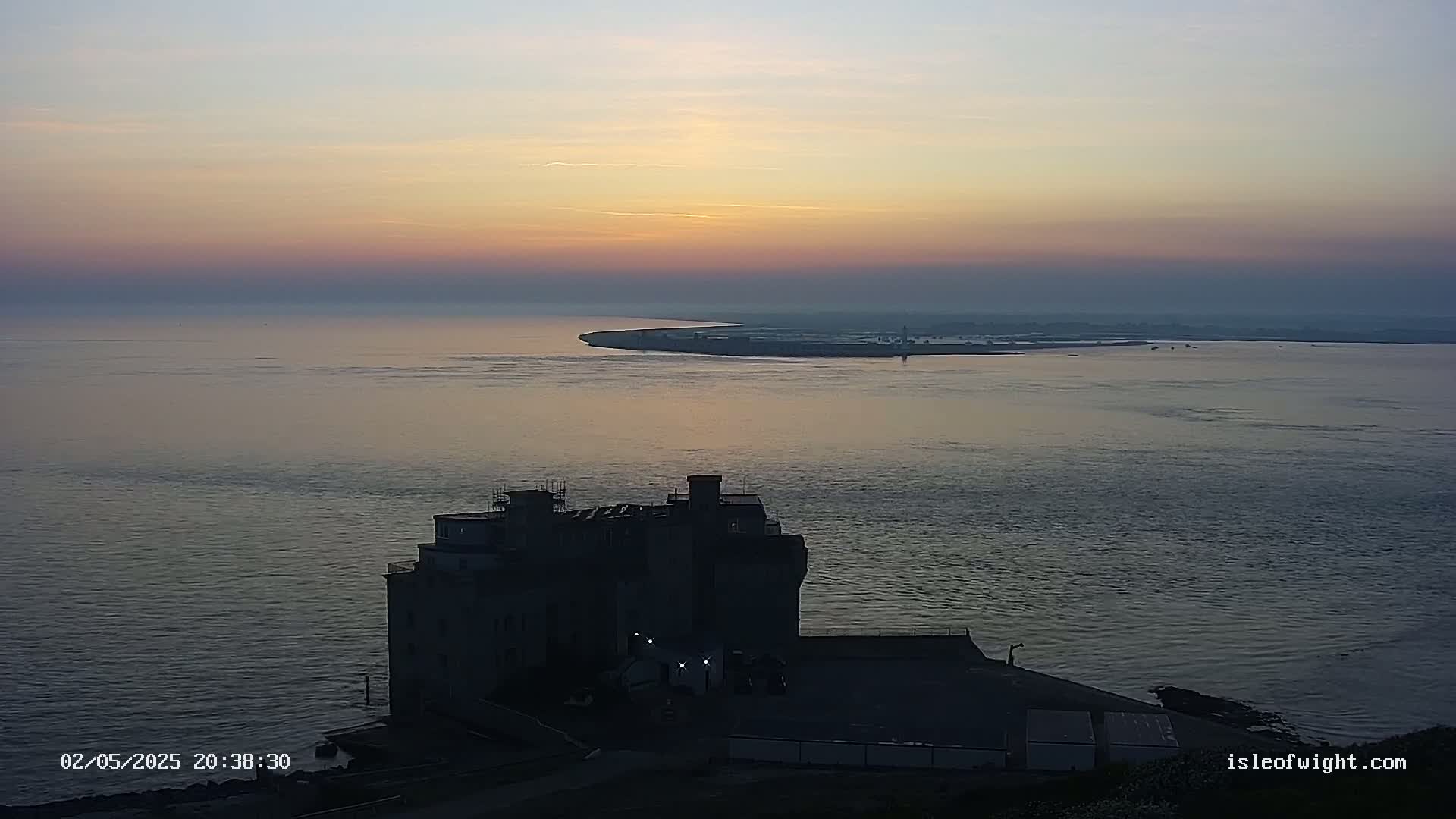 A large, dark building sits on the coast at sunset overlooking calm water and a distant landmass.