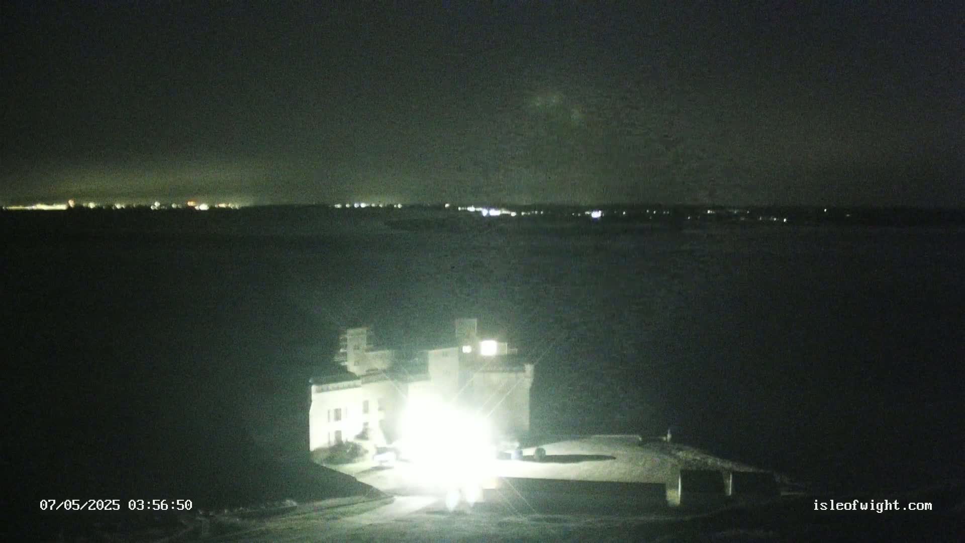 A brightly lit building sits on a dark shore at night, overlooking a body of water with a distant city skyline faintly visible under a clear, dark sky.