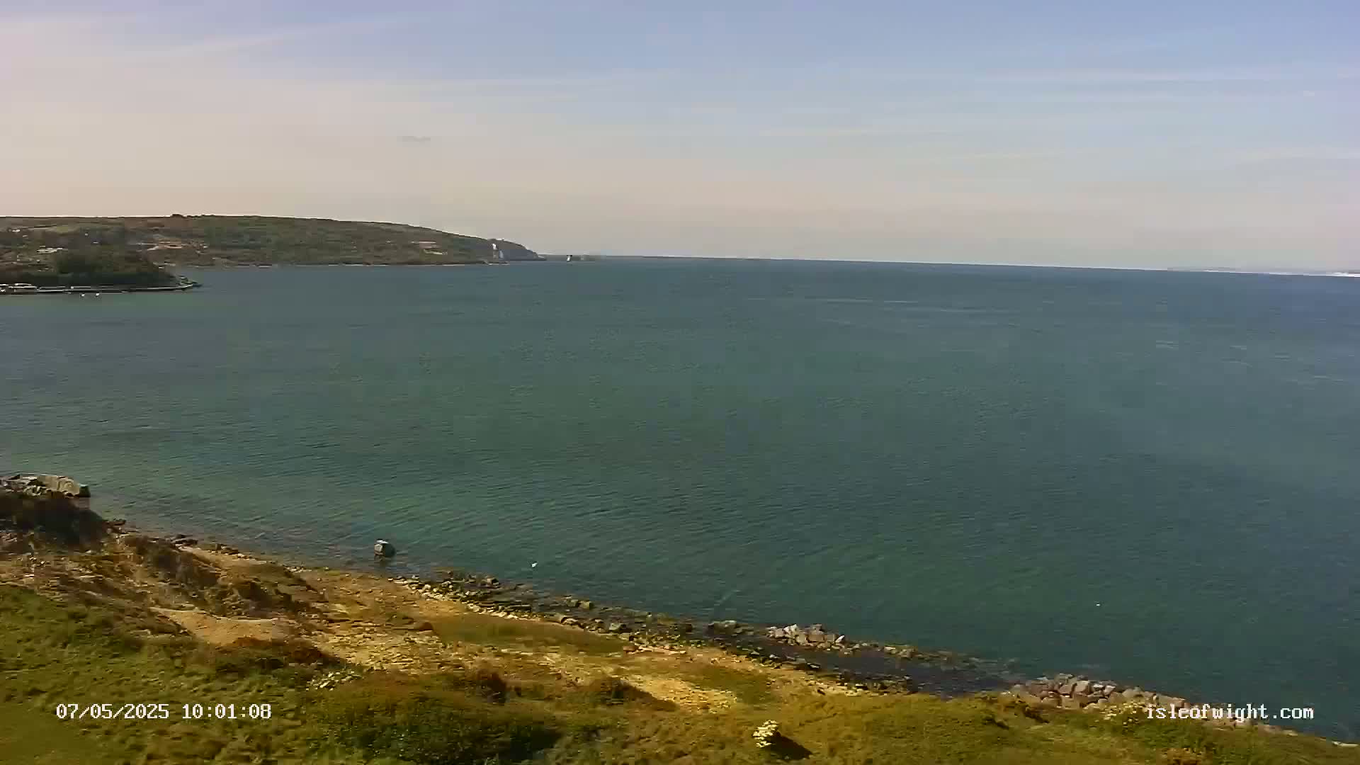 A tranquil sea scene on a sunny day shows a body of water extending to a distant shoreline, with a rocky, grassy slope in the foreground.