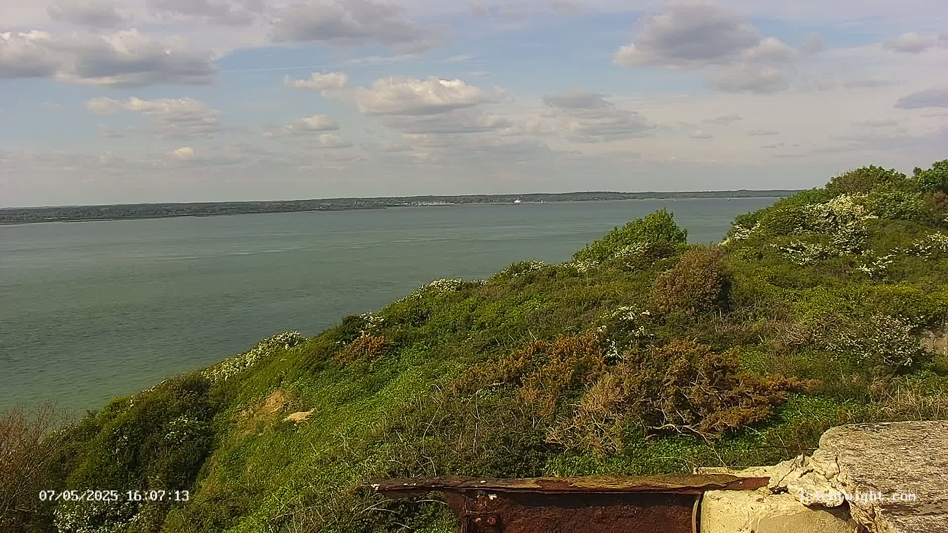 A partly cloudy day reveals a tranquil expanse of water, overlooked by a verdant hillside dotted with flowering shrubs.