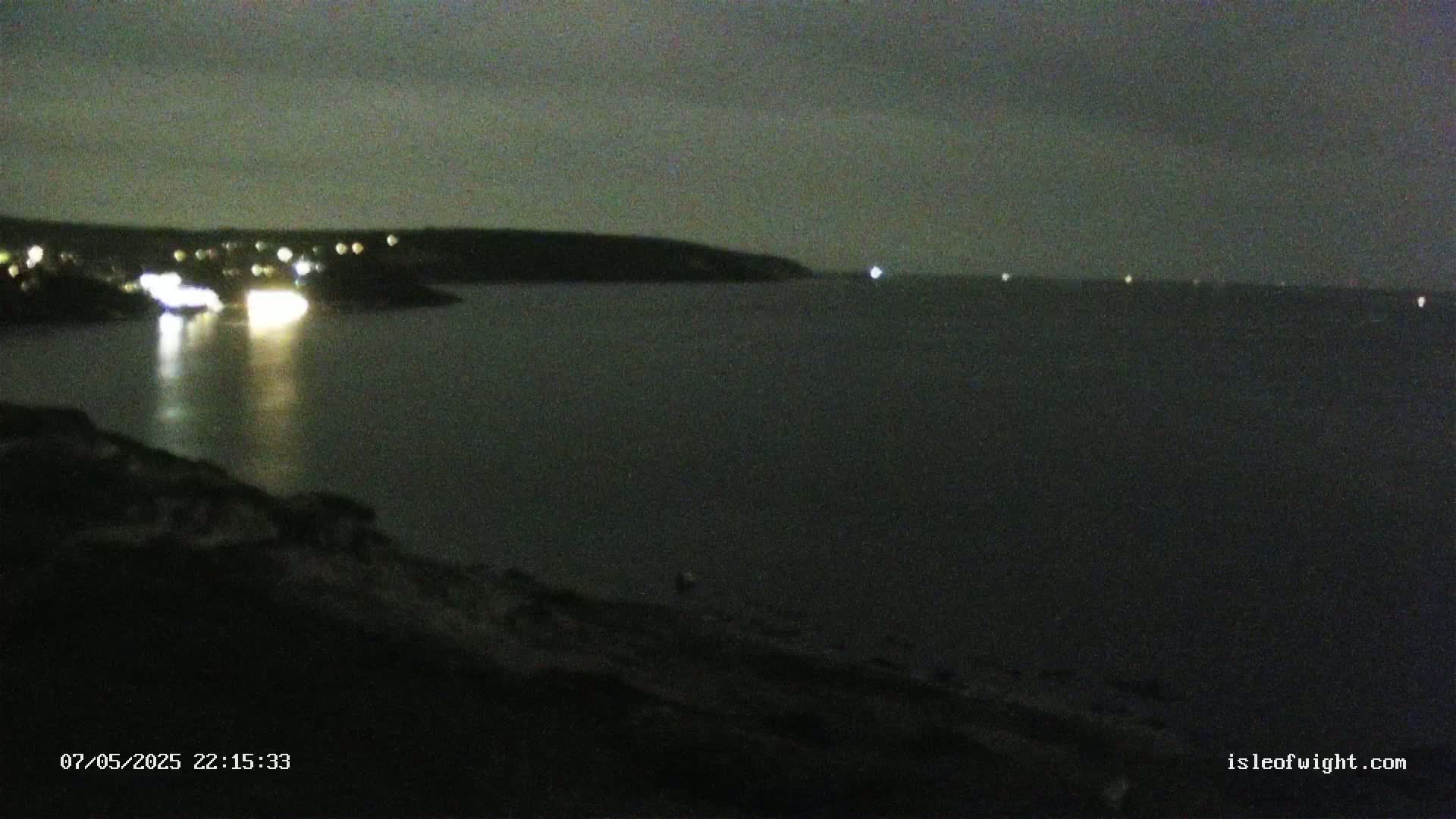 A dark, nighttime scene shows a body of water with lights from a vessel and distant shore lights visible under a mostly clear, dark sky.