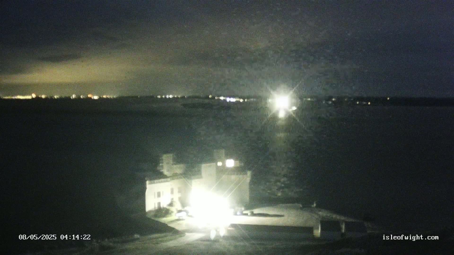 A nighttime view of a light-colored building near a body of water, with a distant city skyline faintly visible under a dark, possibly cloudy sky.