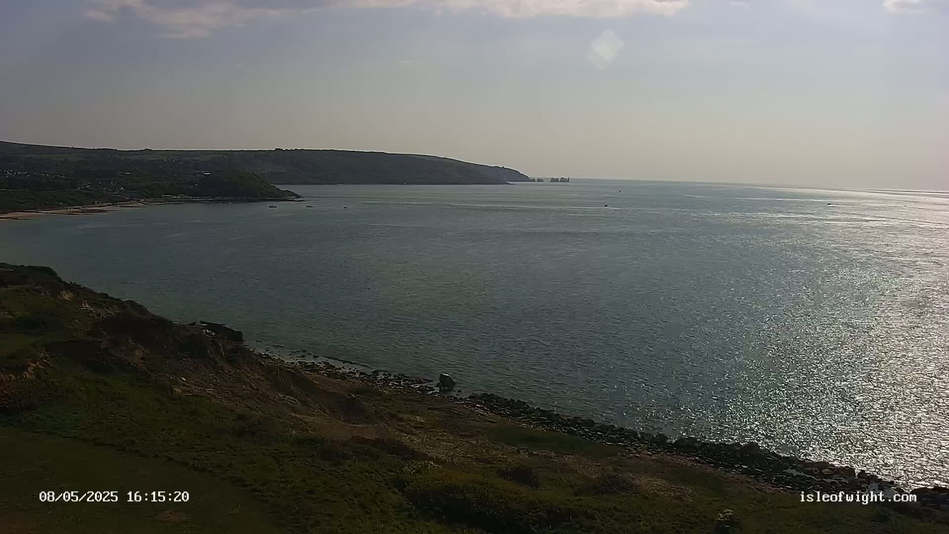 A partly sunny day reveals a calm body of water, stretching from a grassy cliff to a distant coastline, with a few small boats visible on the water.
