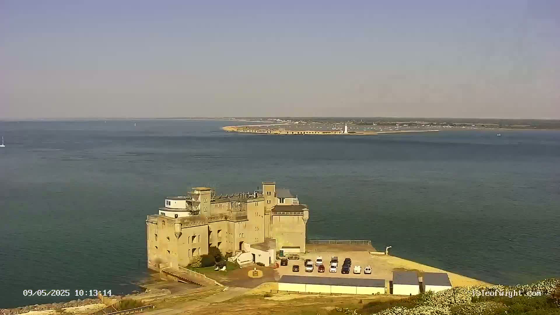 A stone coastal fortress with a parking lot sits on a hill overlooking a calm body of water, with a distant landmass visible on a clear day.