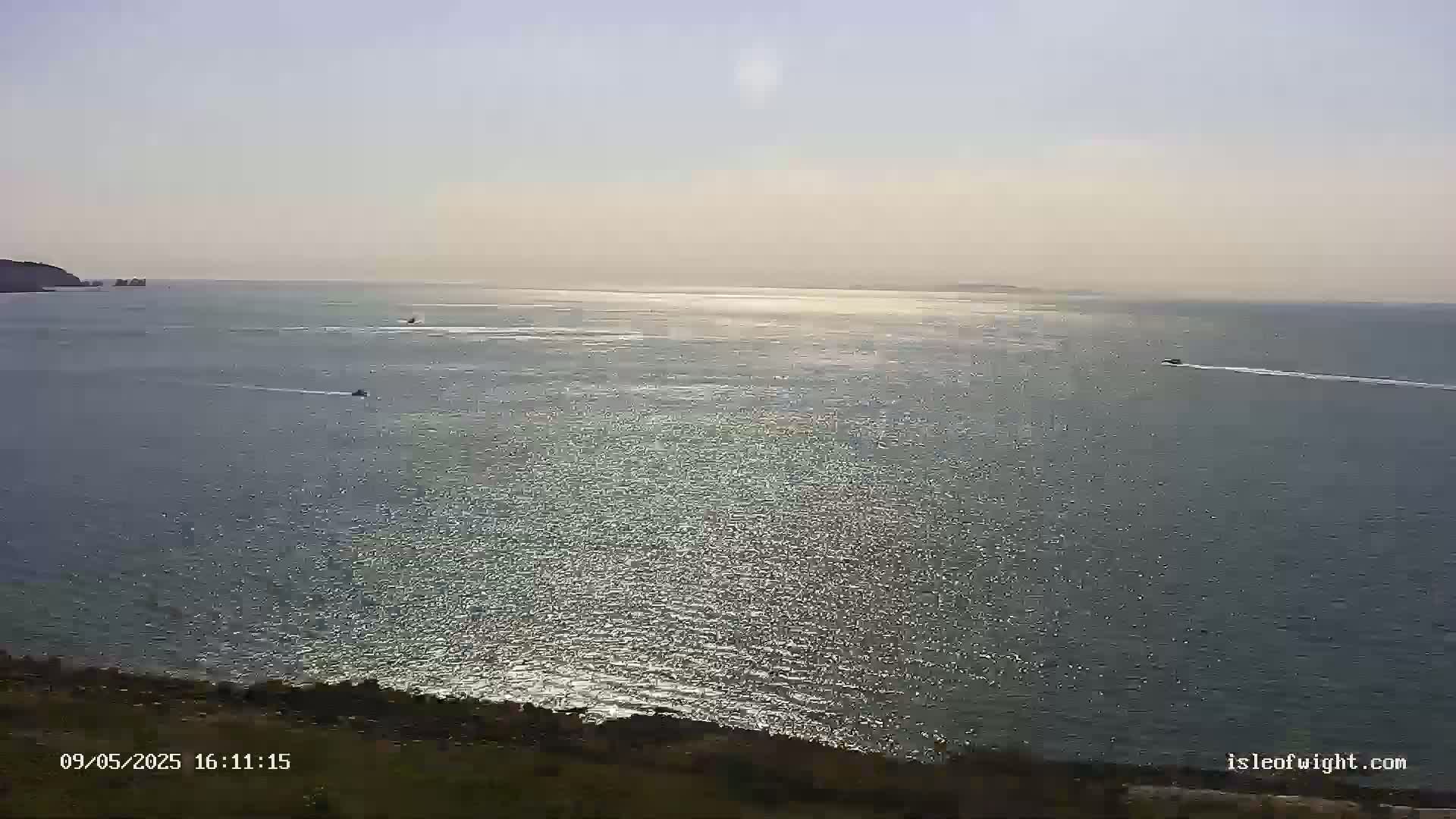 A mostly sunny, calm sea is seen from an elevated vantage point, with several small vessels visible on the water and land in the foreground.