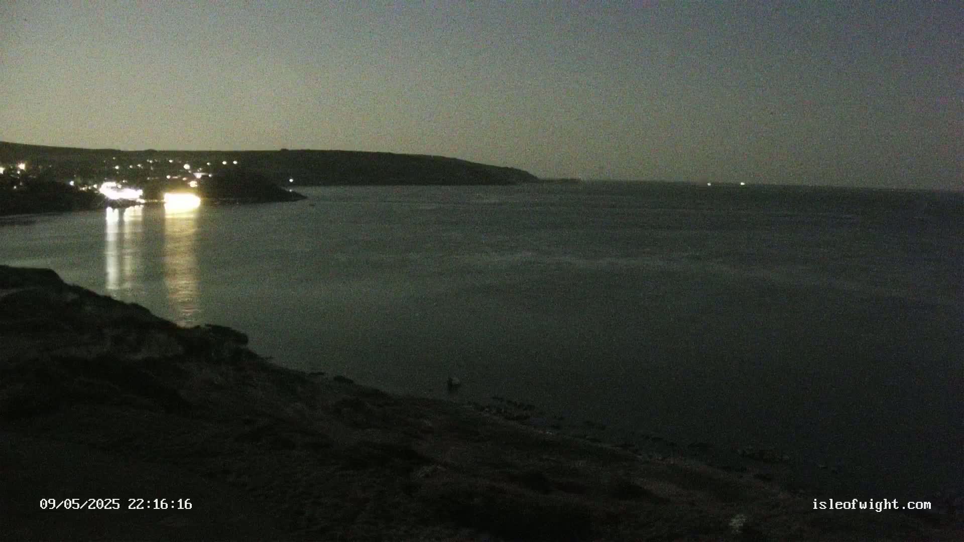 A nighttime coastal scene shows a dark body of water reflecting lights from a town on the shore, under a dark, overcast sky.