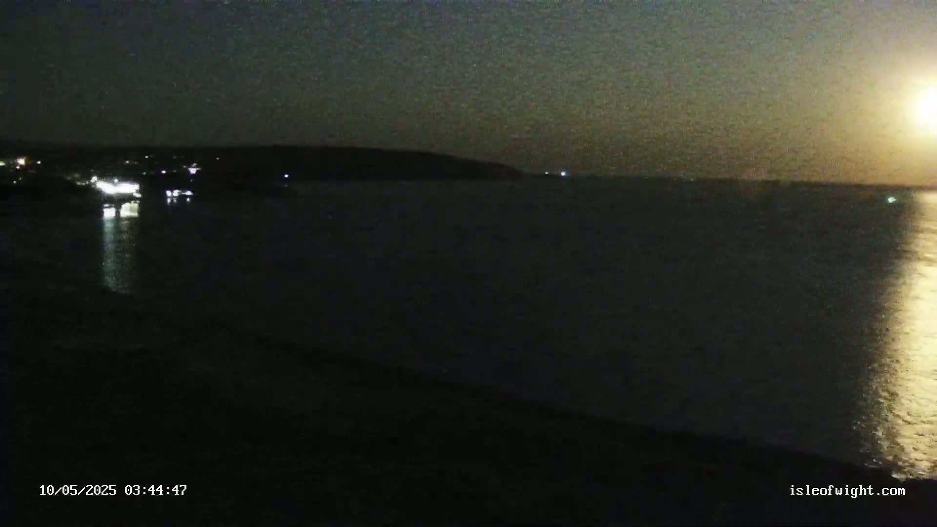 A nighttime view of a dark body of water reflecting a bright moon, with a dark shoreline and faintly lit landmass in the distance.