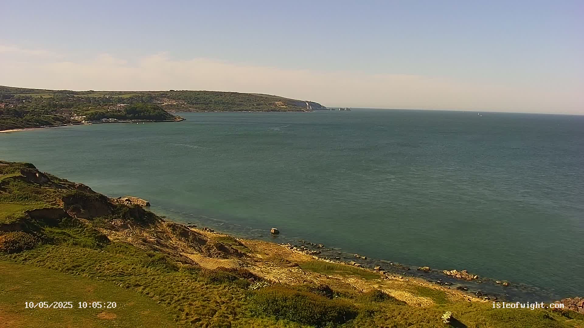 A grassy cliff overlooks a calm, green sea on a sunny day, with a distant coastline and a few small boats visible.