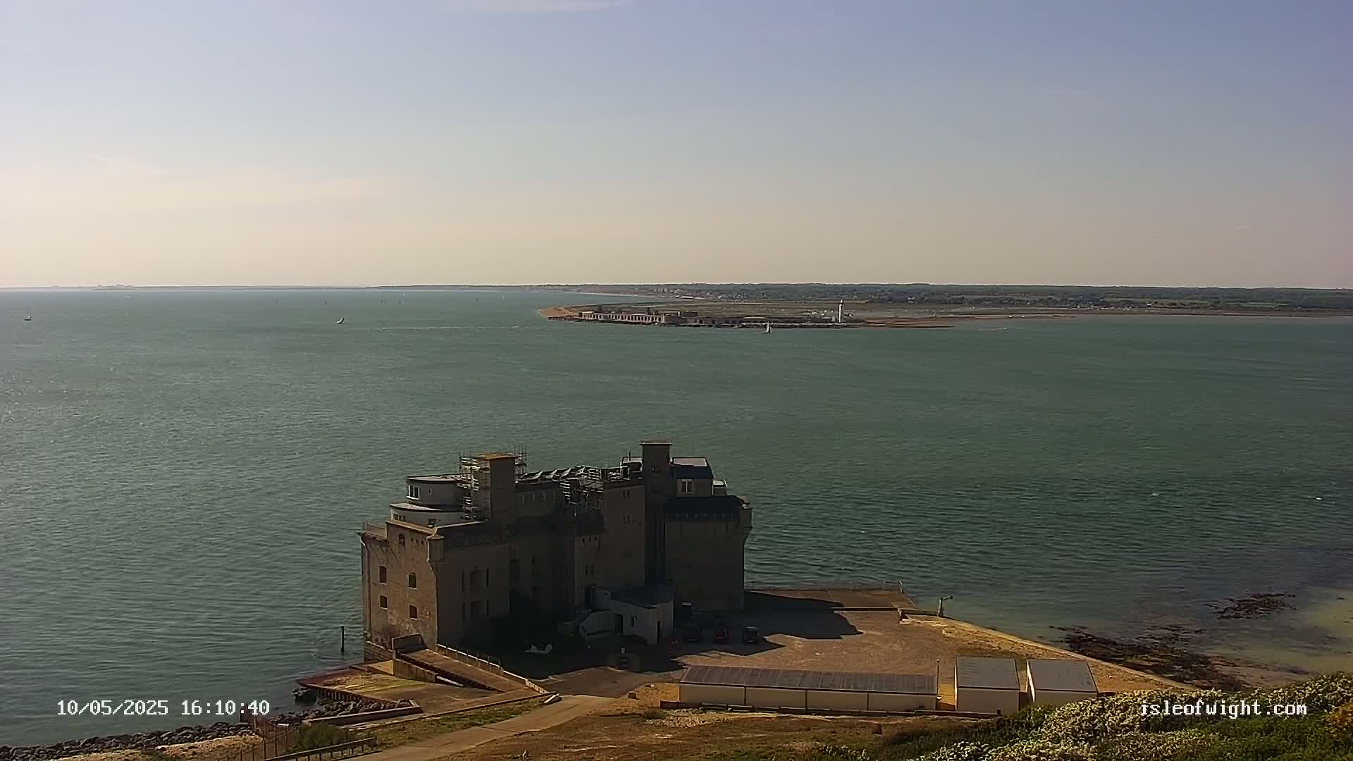 A stone castle-like structure sits on a coast overlooking a calm body of water under a clear sky.