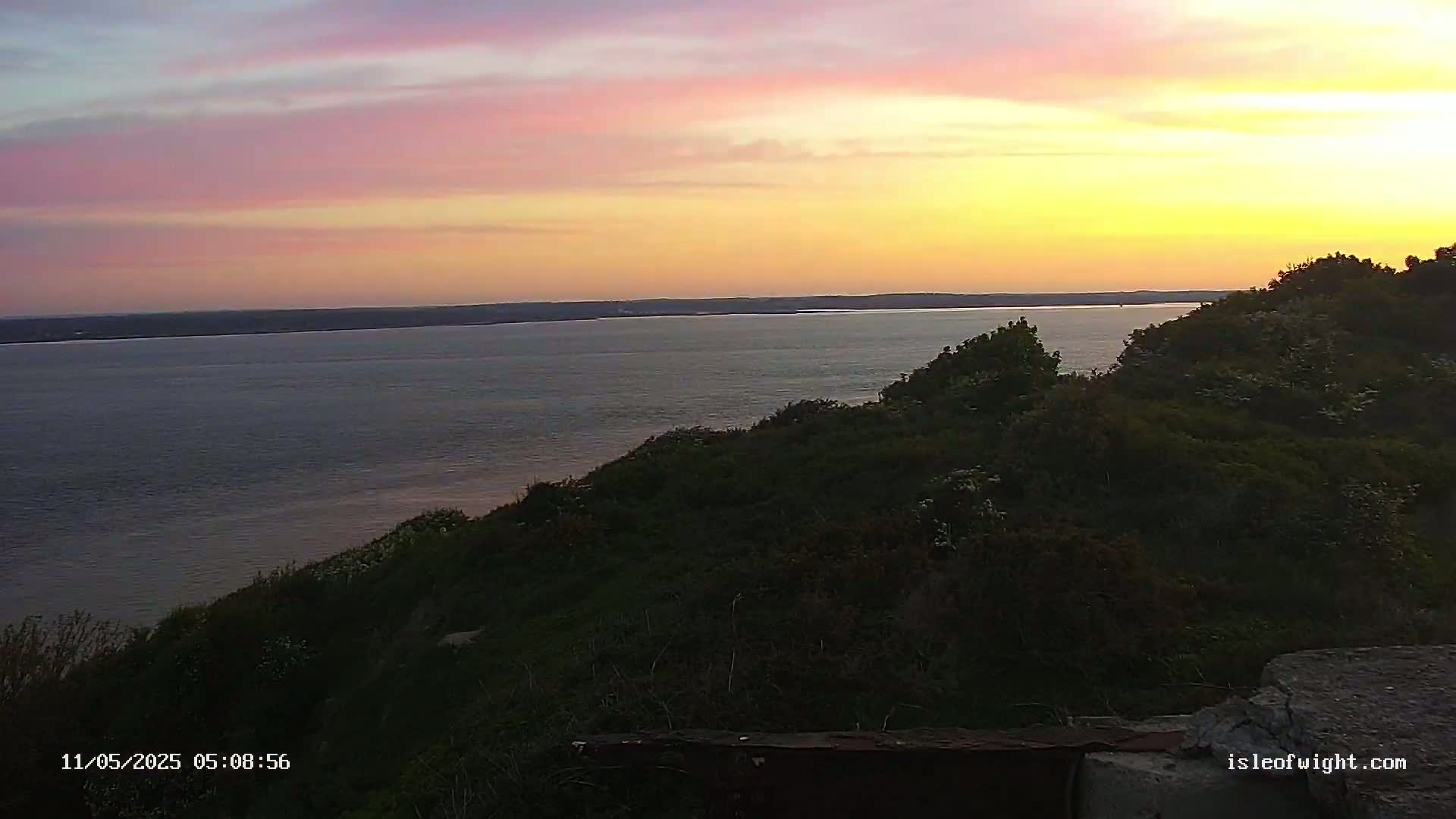 A calm body of water stretches to the horizon under a sunset sky of pink and yellow hues, viewed from a verdant hillside.
