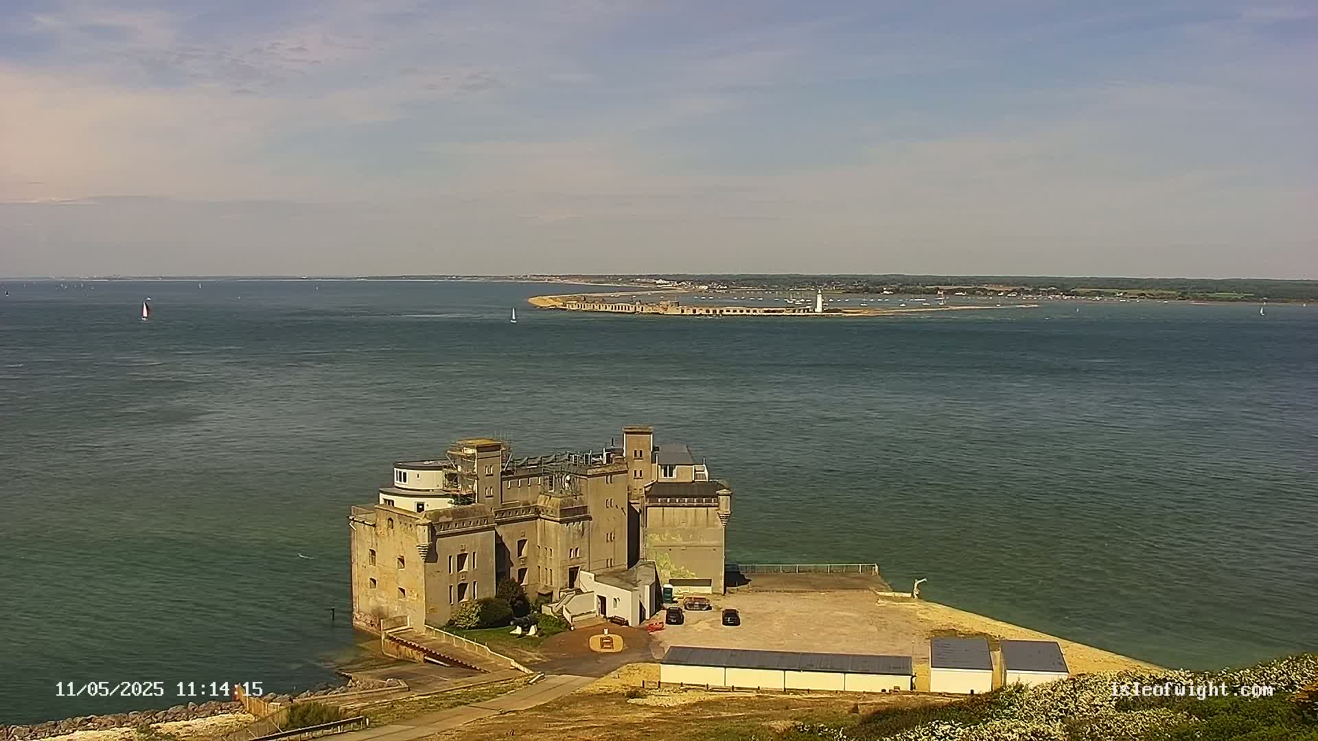A large stone structure sits on a coast overlooking a calm body of water on a partly cloudy day, with a distant landmass visible in the background.