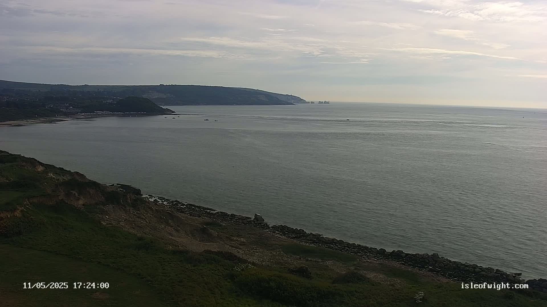 A view of a calm sea with a coastline featuring cliffs and green land, under a mostly cloudy sky.