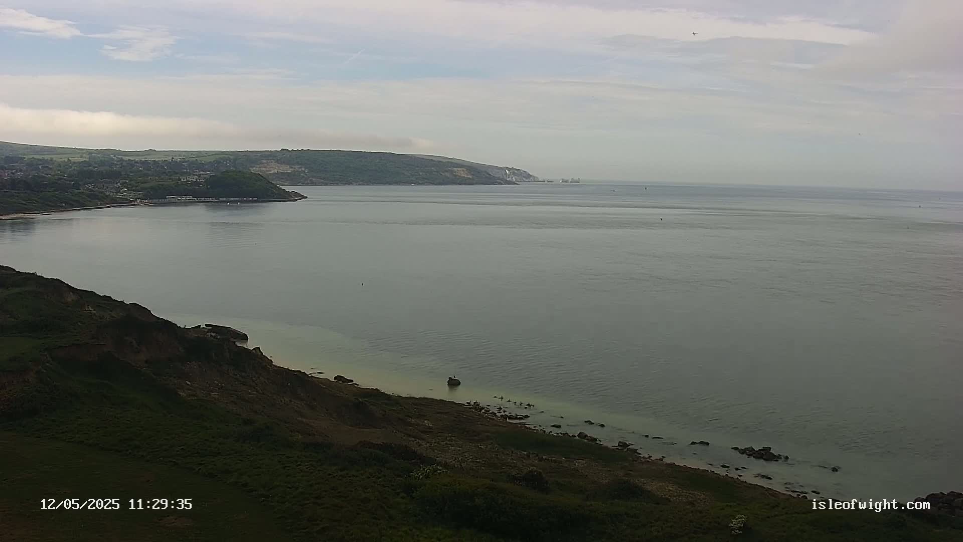 A calm, overcast day shows a wide expanse of sea separating a green, hilly coastline from a distant, similar coastline.
