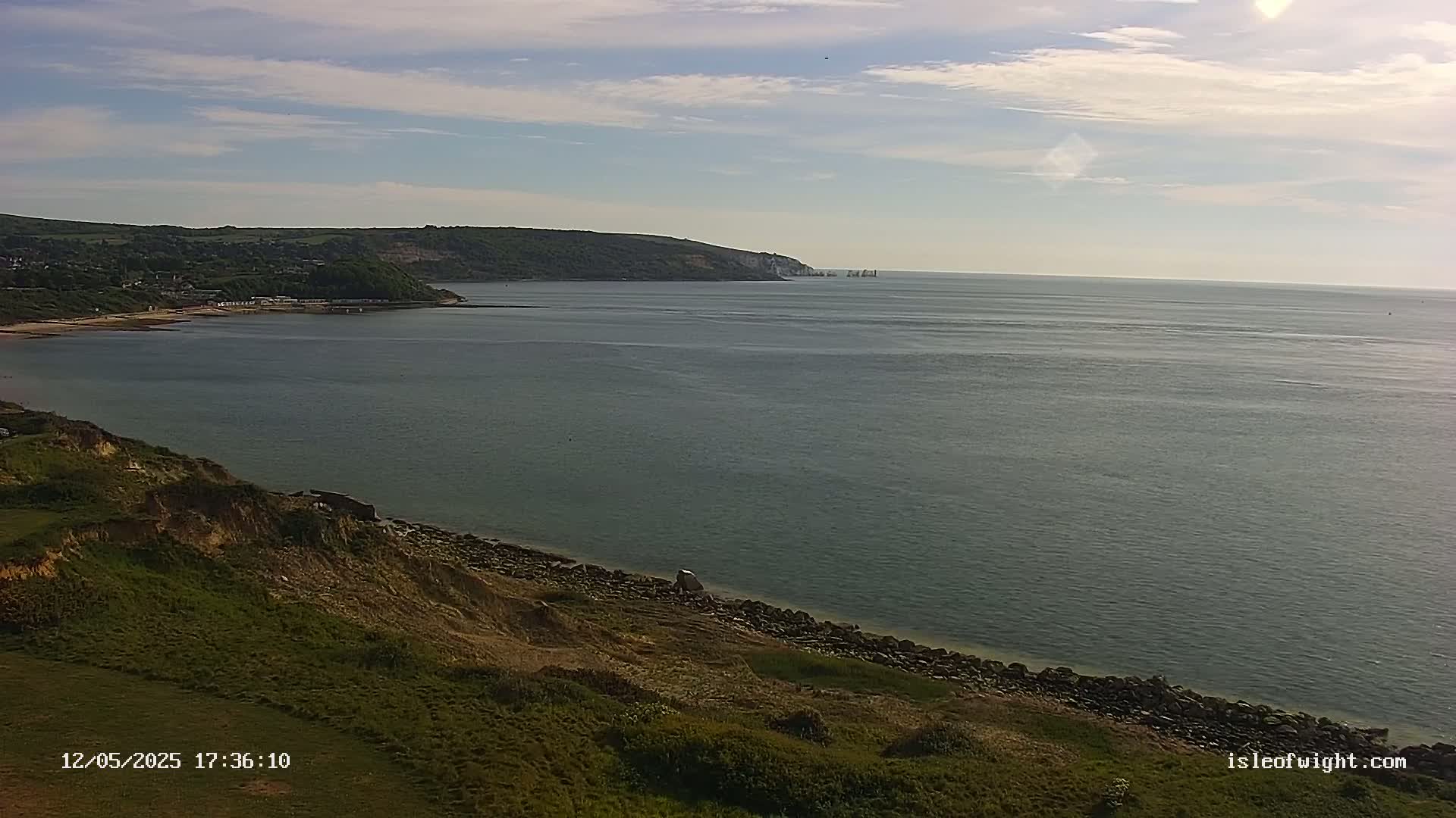 A mostly sunny day reveals a calm sea lapping a rocky shoreline, with green hills and distant land visible in the background.