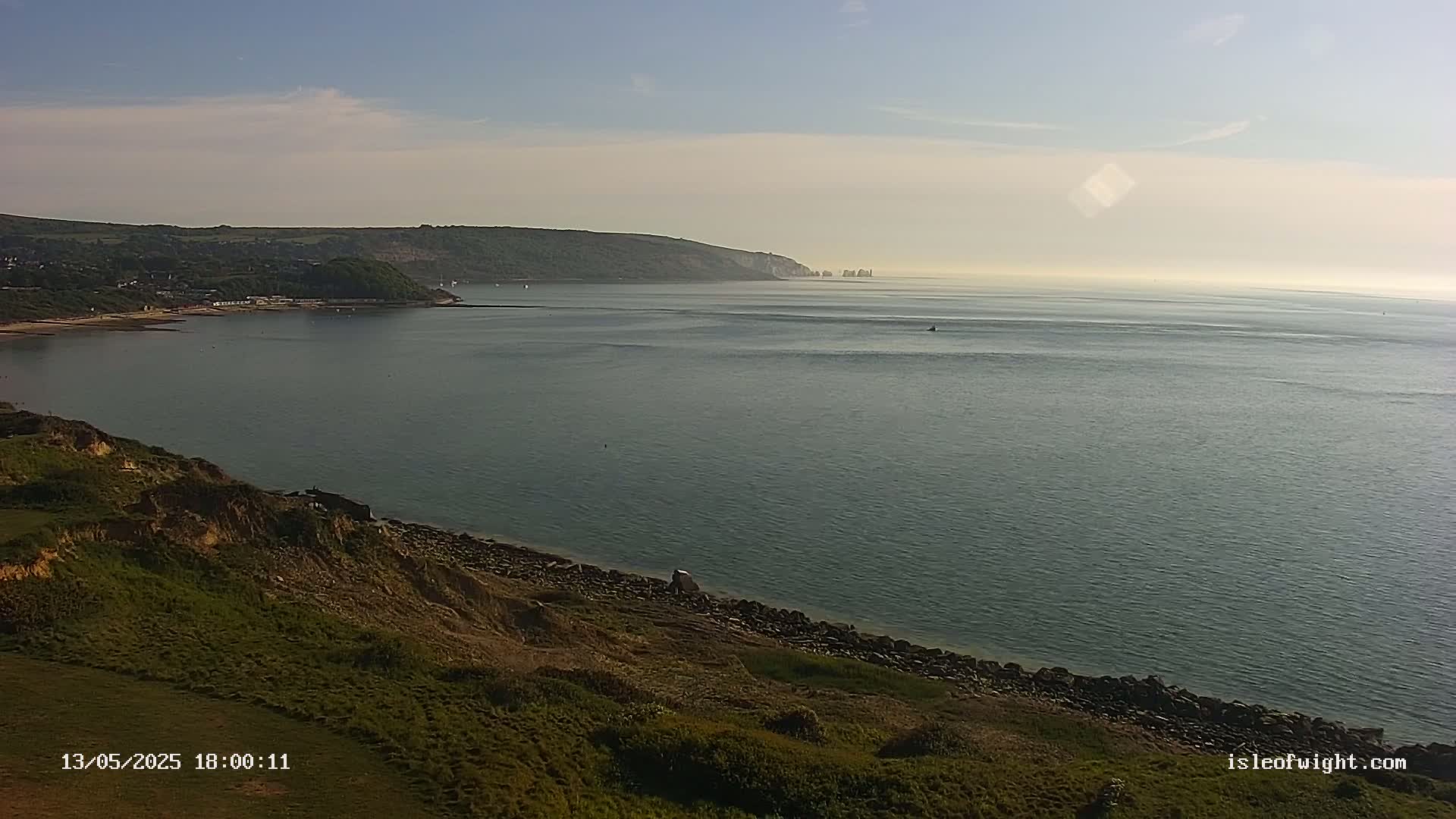 A calm, expansive body of water stretches to a distant coastline under a mostly clear sky, with grassy hillsides and a rocky shoreline in the foreground.