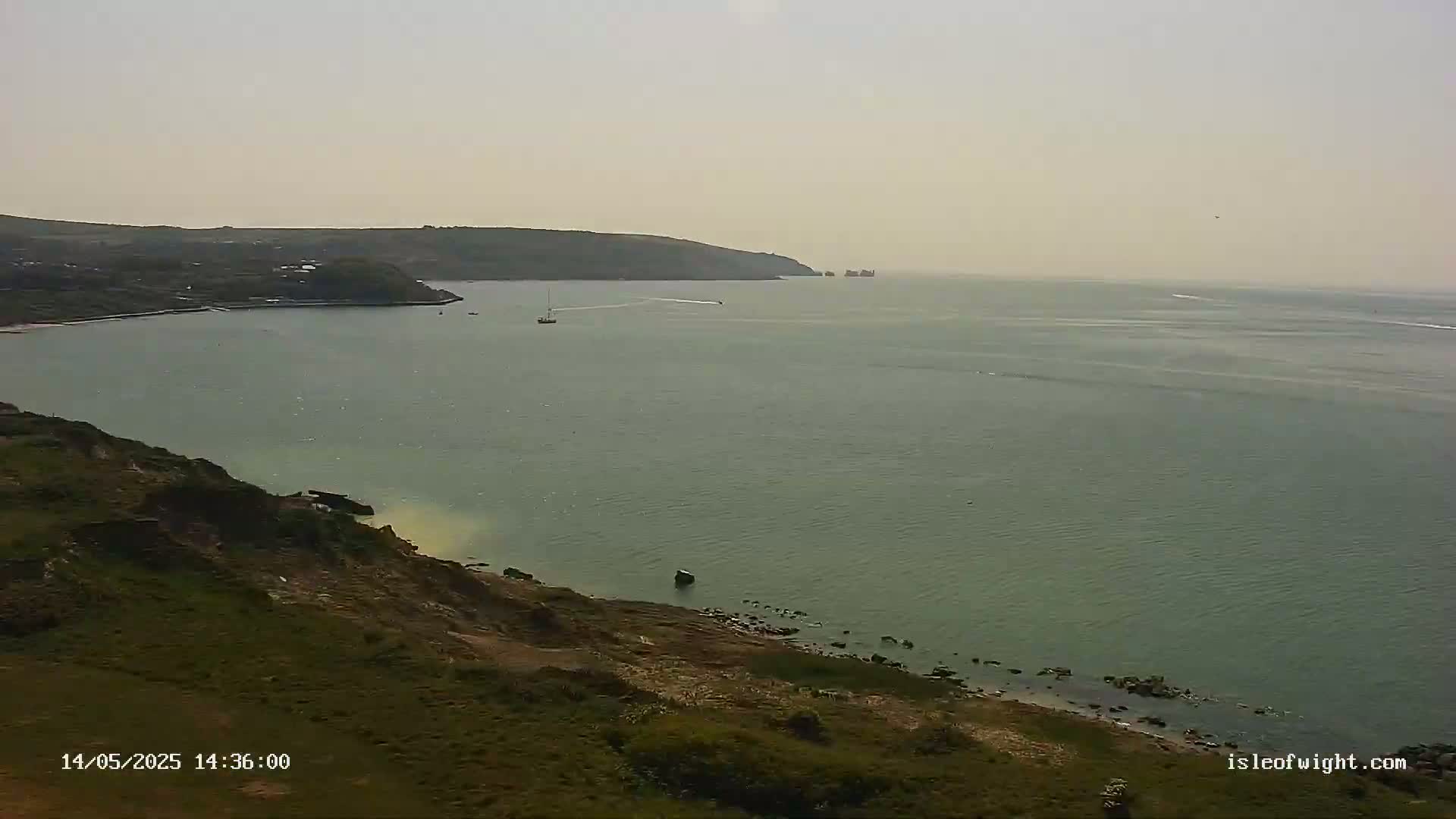 A hazy, sunny day overlooks a calm bay with a few boats, a grassy cliff, and distant land.