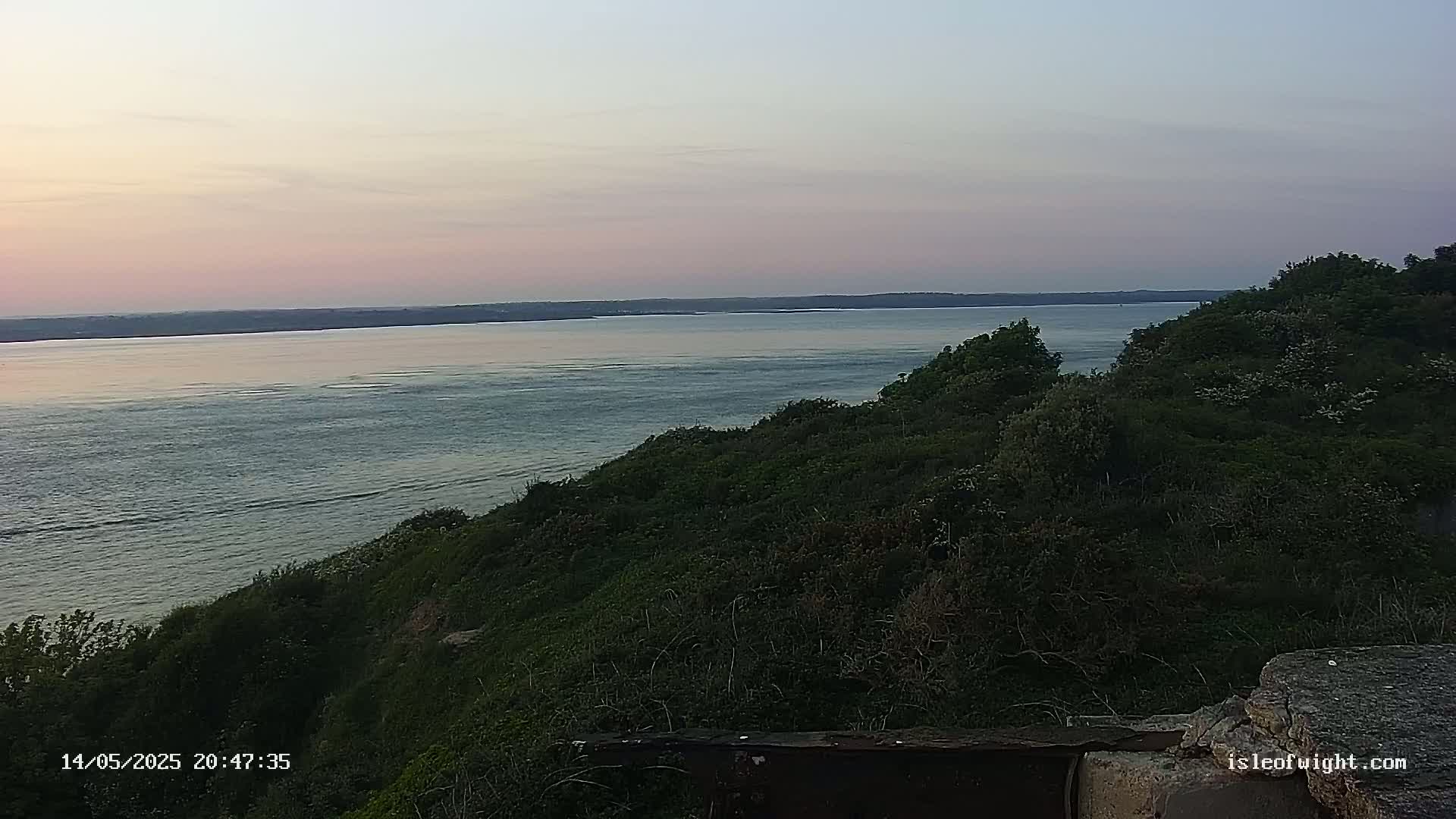 A tranquil coastal scene at sunset, showing a calm body of water stretching to a distant landmass, overlooked by a verdant hillside.