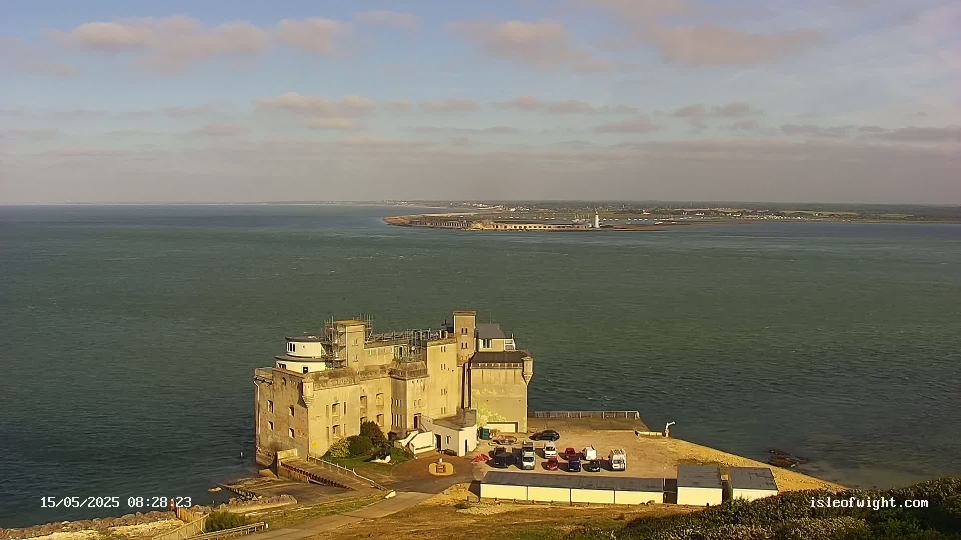 A large stone building, possibly a castle or fort, sits on a coast overlooking a calm body of water under a partly cloudy sky.