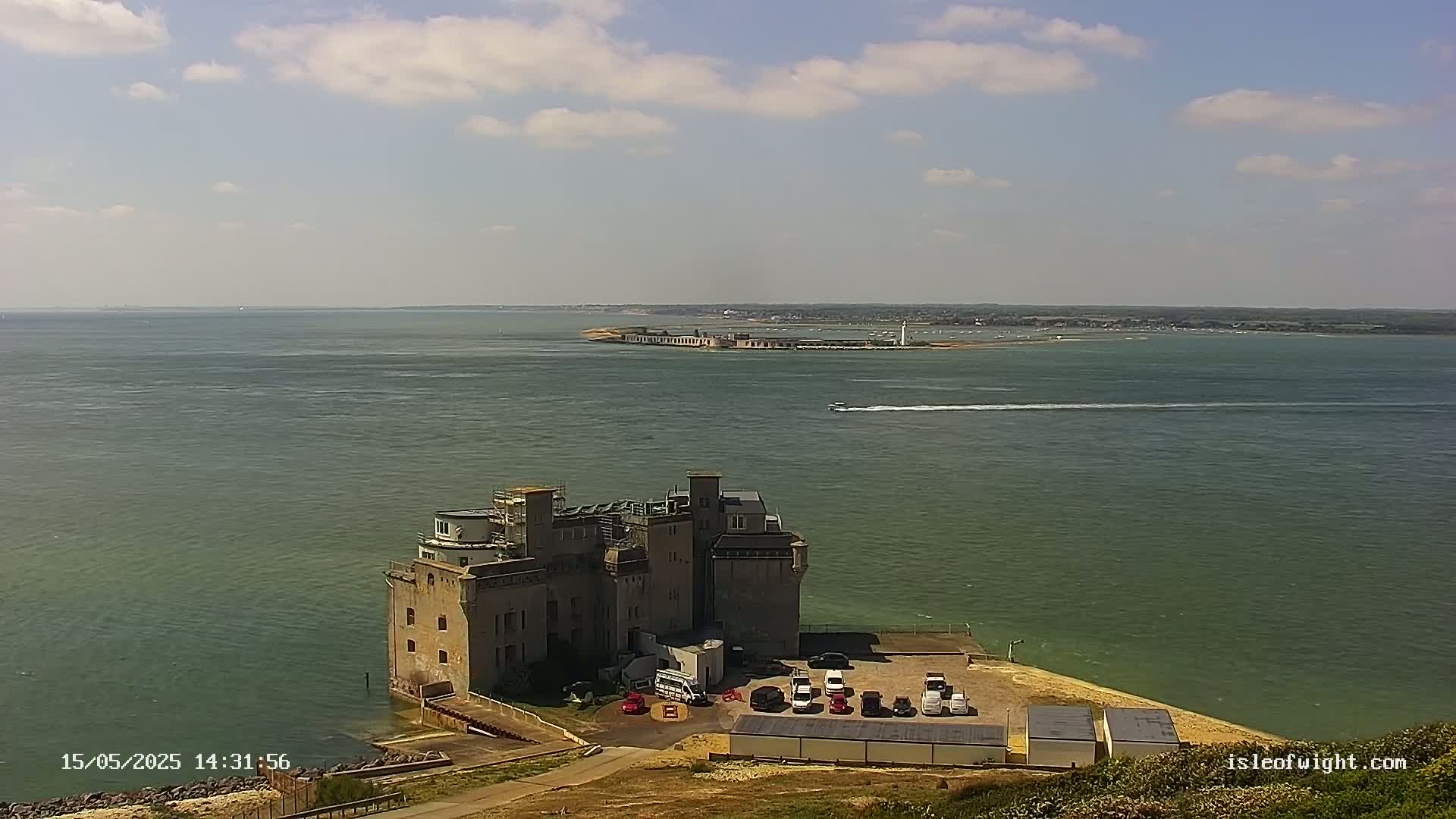 A stone fortress-like building sits on a coastal point overlooking a calm body of water under a partly cloudy sky.