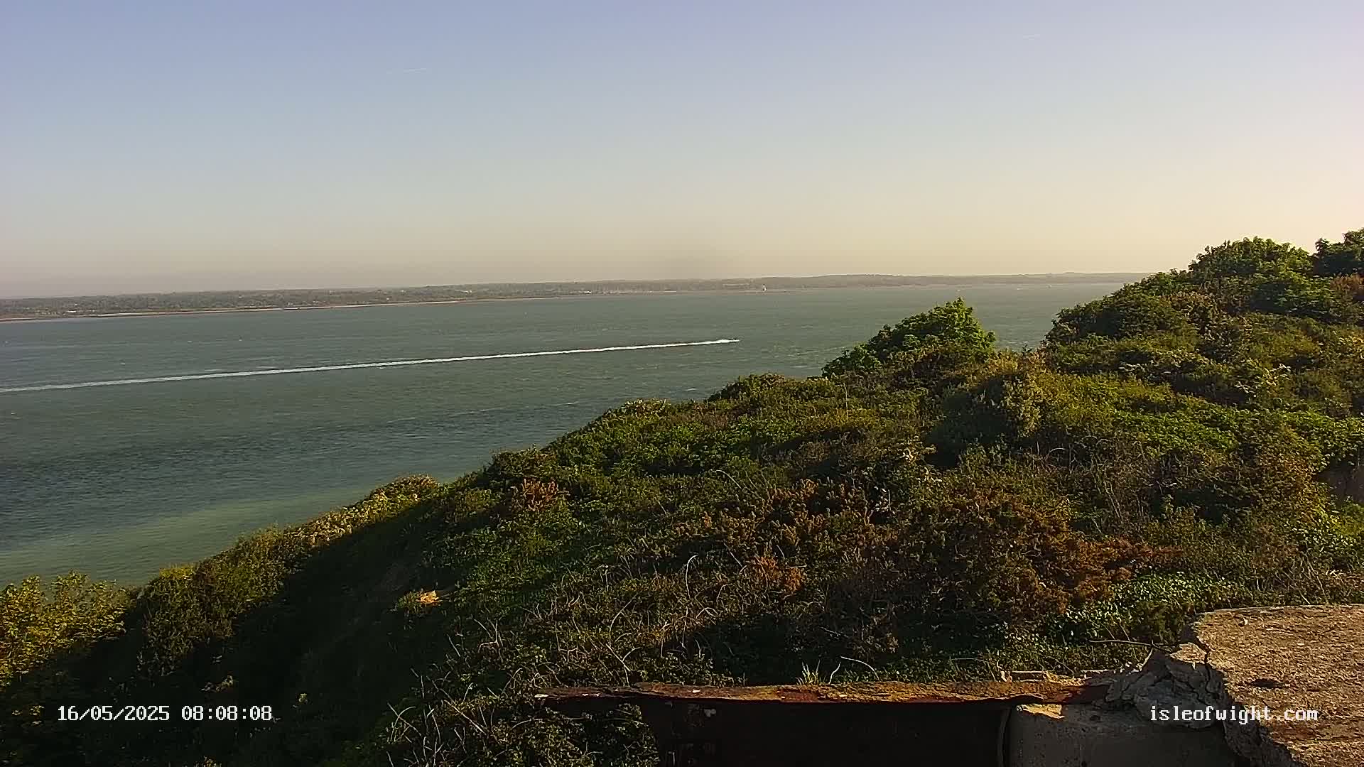 A boat's wake is visible on a calm body of water viewed from a green, hilly coastal overlook under a clear sky.