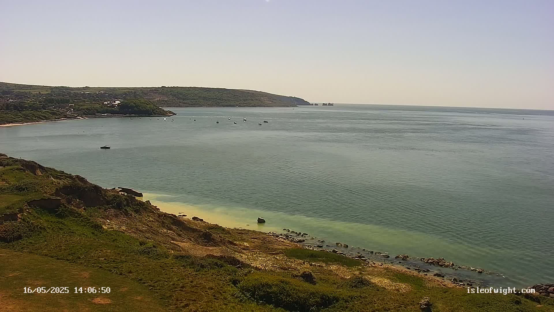 A calm, sunny day reveals a coastal scene with a bay, a grassy hillside, and a few boats scattered on the water.