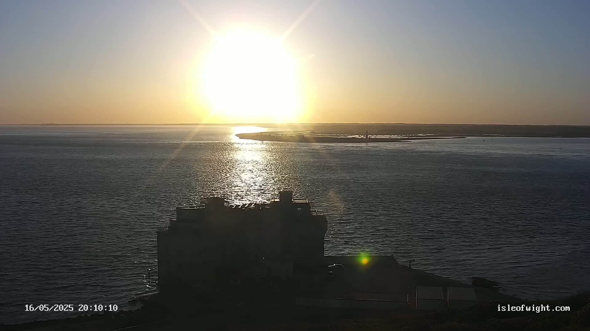 A bright sunset over calm water, with a building silhouetted in the foreground.
