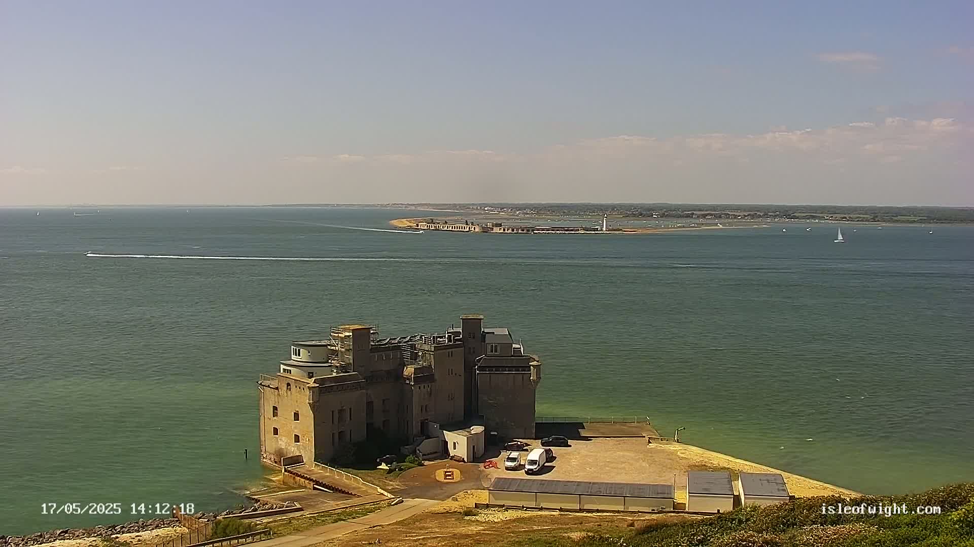 A stone fortress sits on a coastal point overlooking a calm body of water with a distant landmass under a partly cloudy sky.