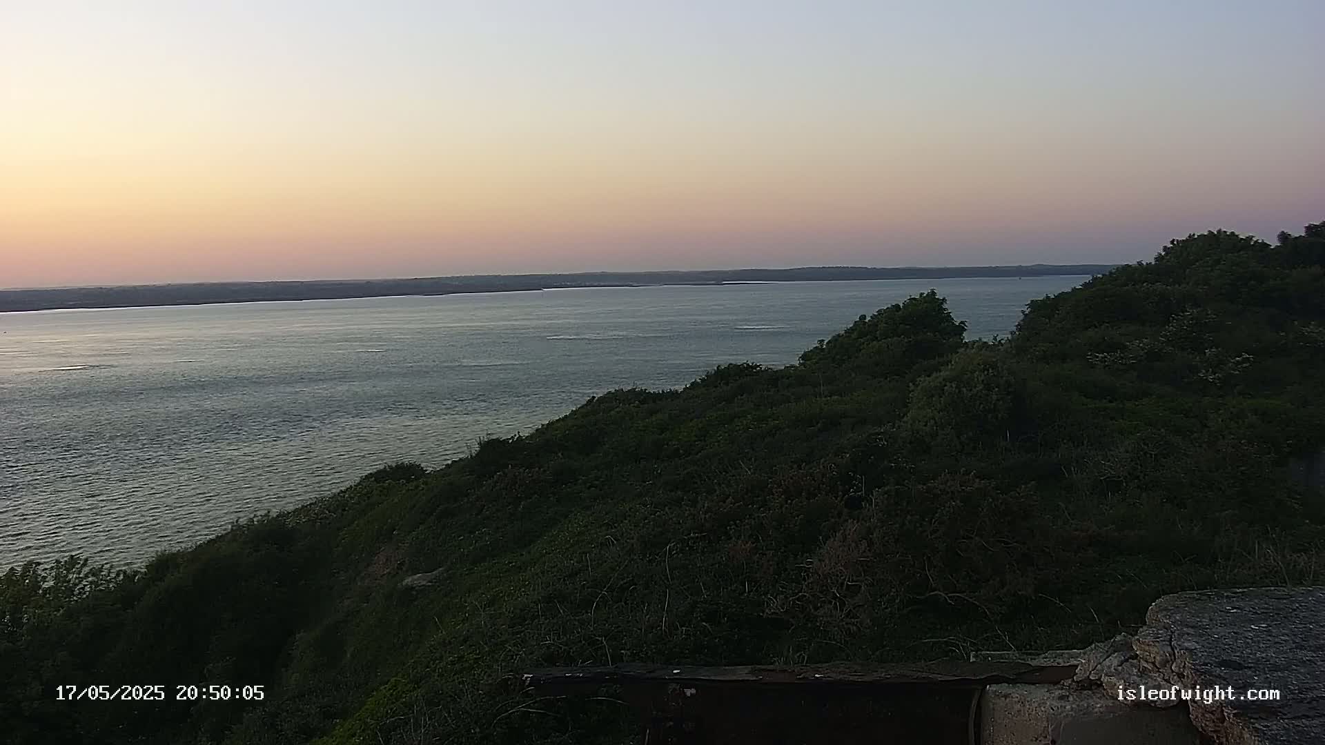 A calm body of water stretches to the horizon under a pale pink and purple sunset, viewed from a verdant hillside.