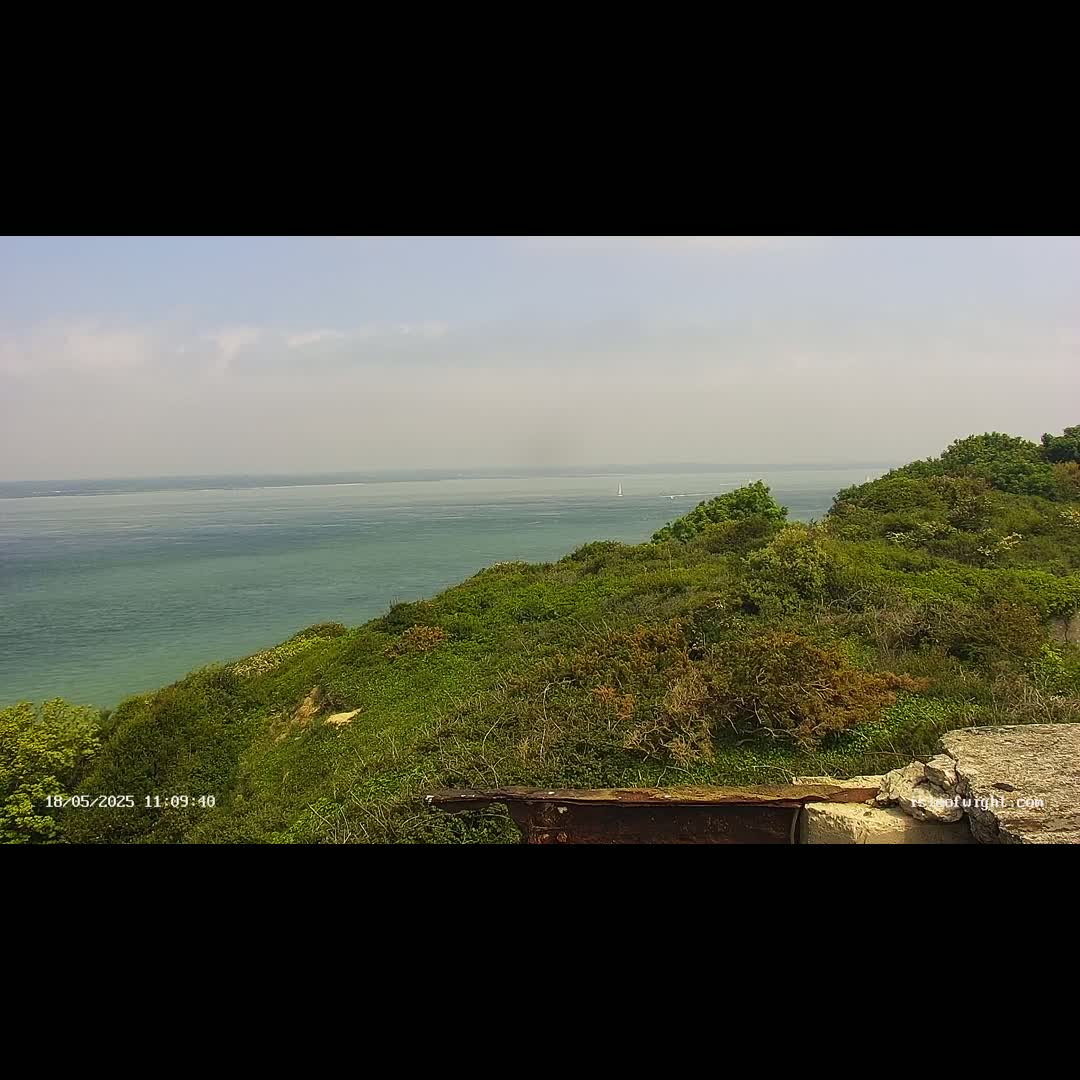 A view from a grassy clifftop overlooking a calm, teal sea on a partly cloudy day.