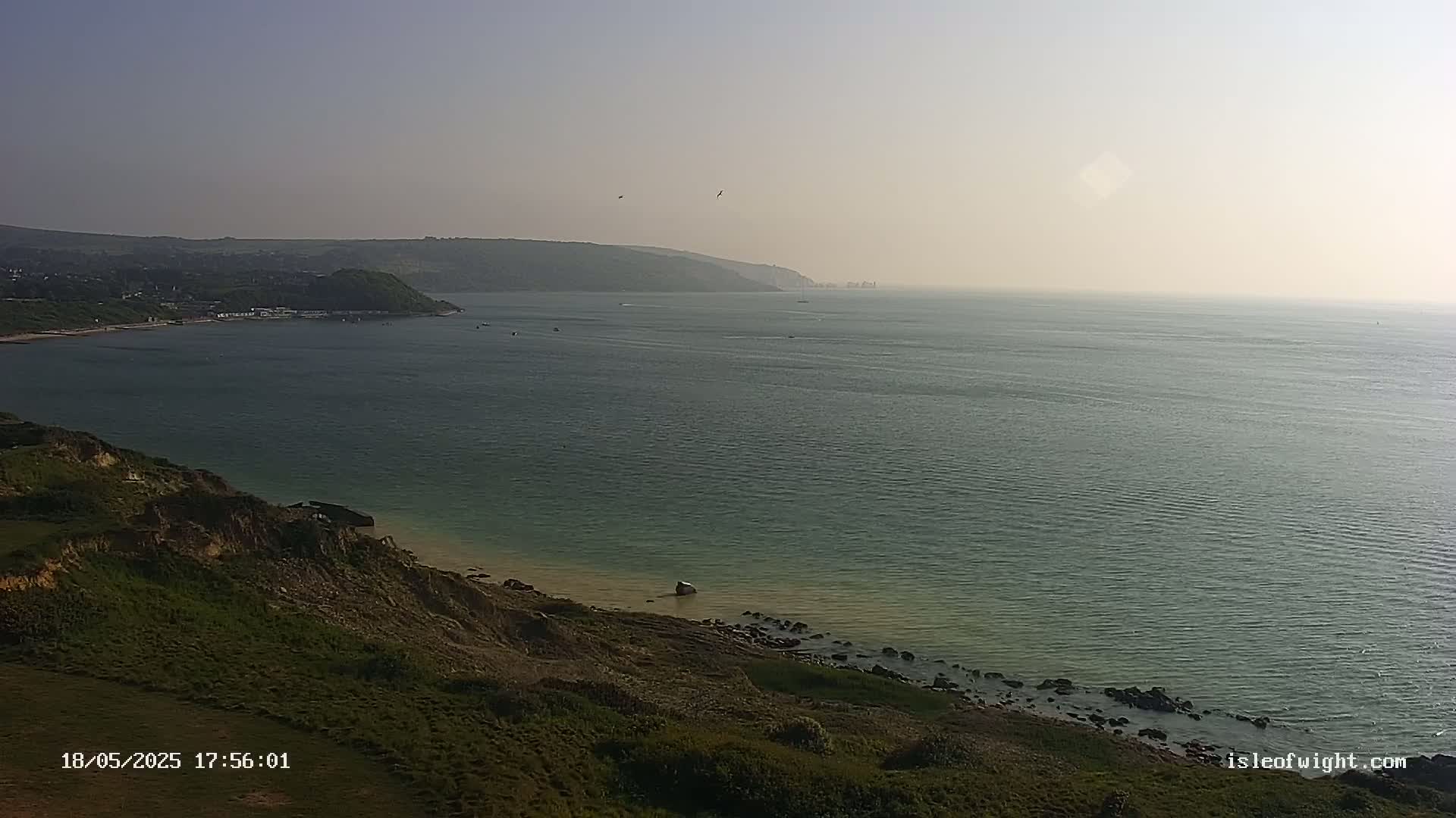 A hazy, sunny day reveals a coastal scene with a calm sea, a rocky shoreline, and green hills in the distance.