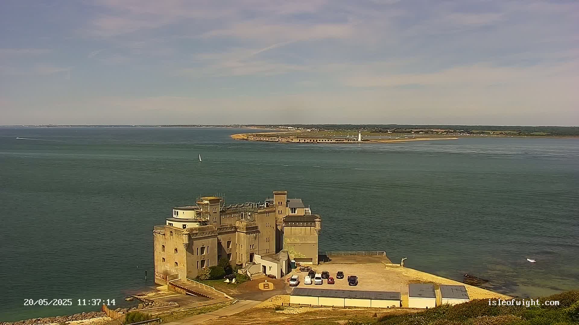 A large stone building sits on a coast overlooking calm, dark-green water under a mostly clear sky.