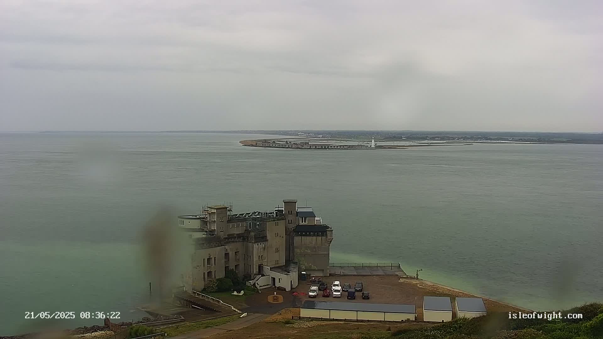 A coastal scene under an overcast sky shows a large building overlooking a body of water with a distant island and lighthouse.